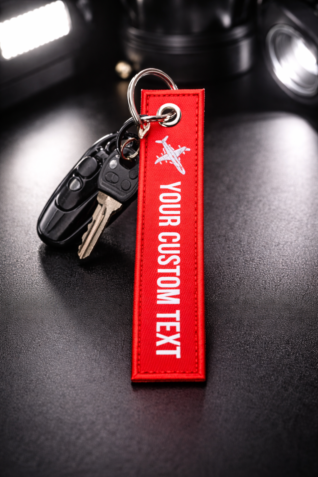 Car key fob with a red luggage tag that says 'Your Custom Text' and an airplane icon, placed on a black surface with a blurred background.