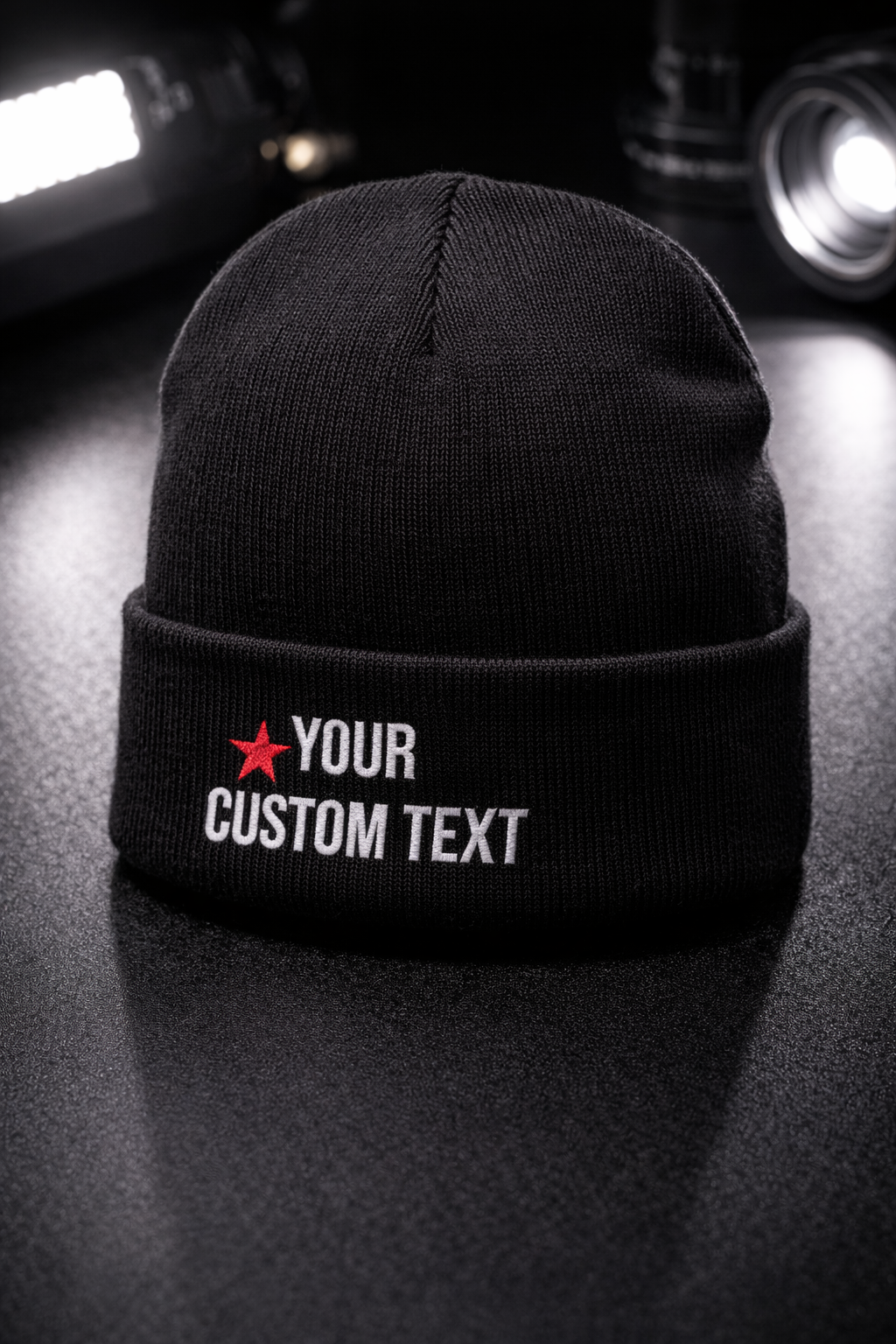Black beanie hat with customizable text, featuring a red star, placed on a dark surface with cameras in the background.
