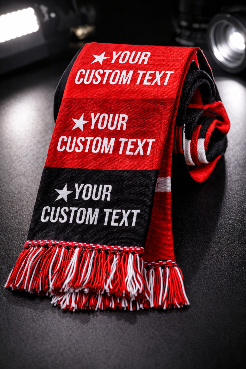A sports scarf with red, black, and white stripes and the words "YOUR CUSTOM TEXT" in white, placed on a dark surface with a DJ setup in the background.