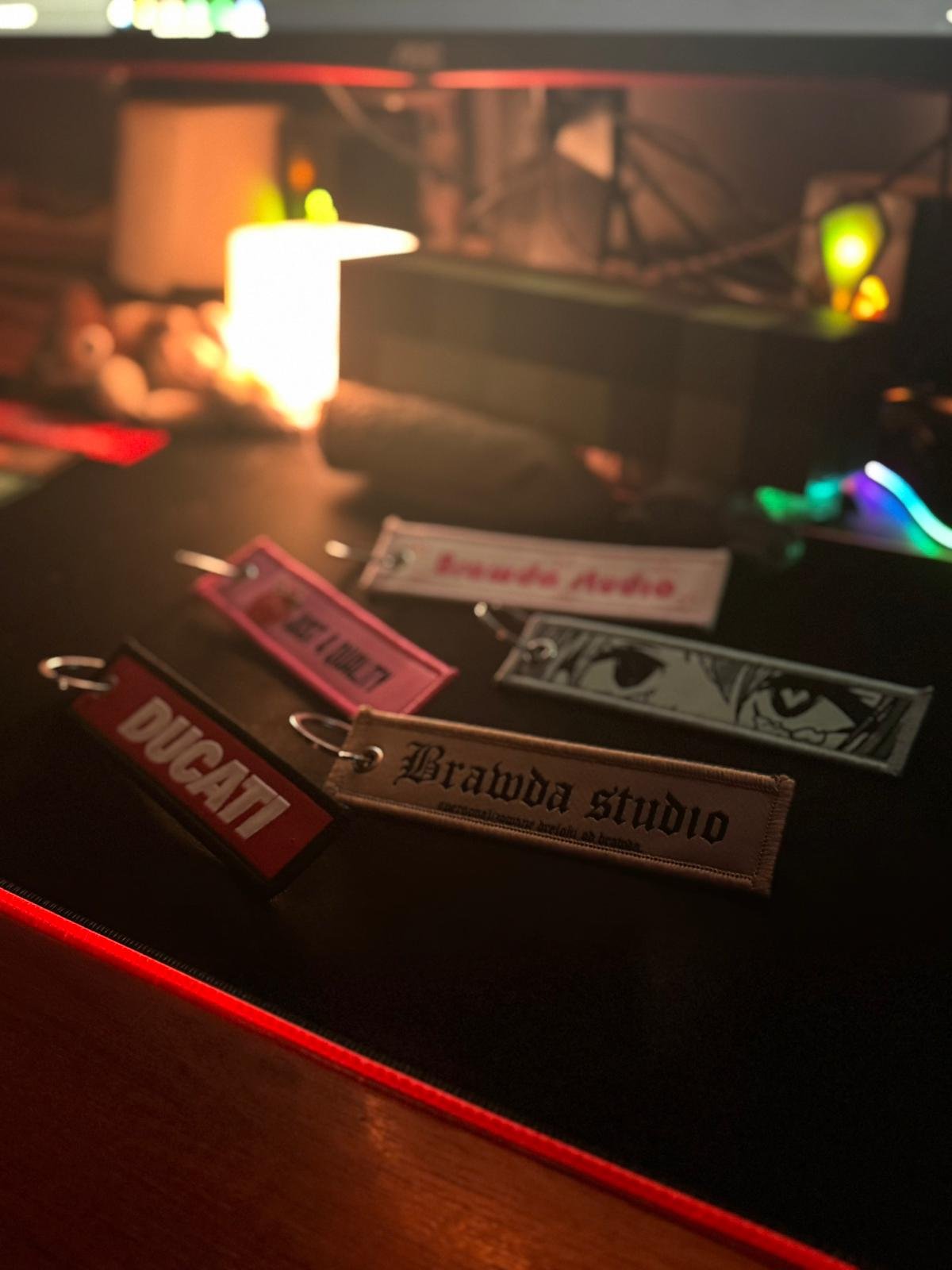 Close-up of multiple fabric keychains with logo and text, on a dark surface in a dimly lit environment, with blurred colorful lights and objects in the background.