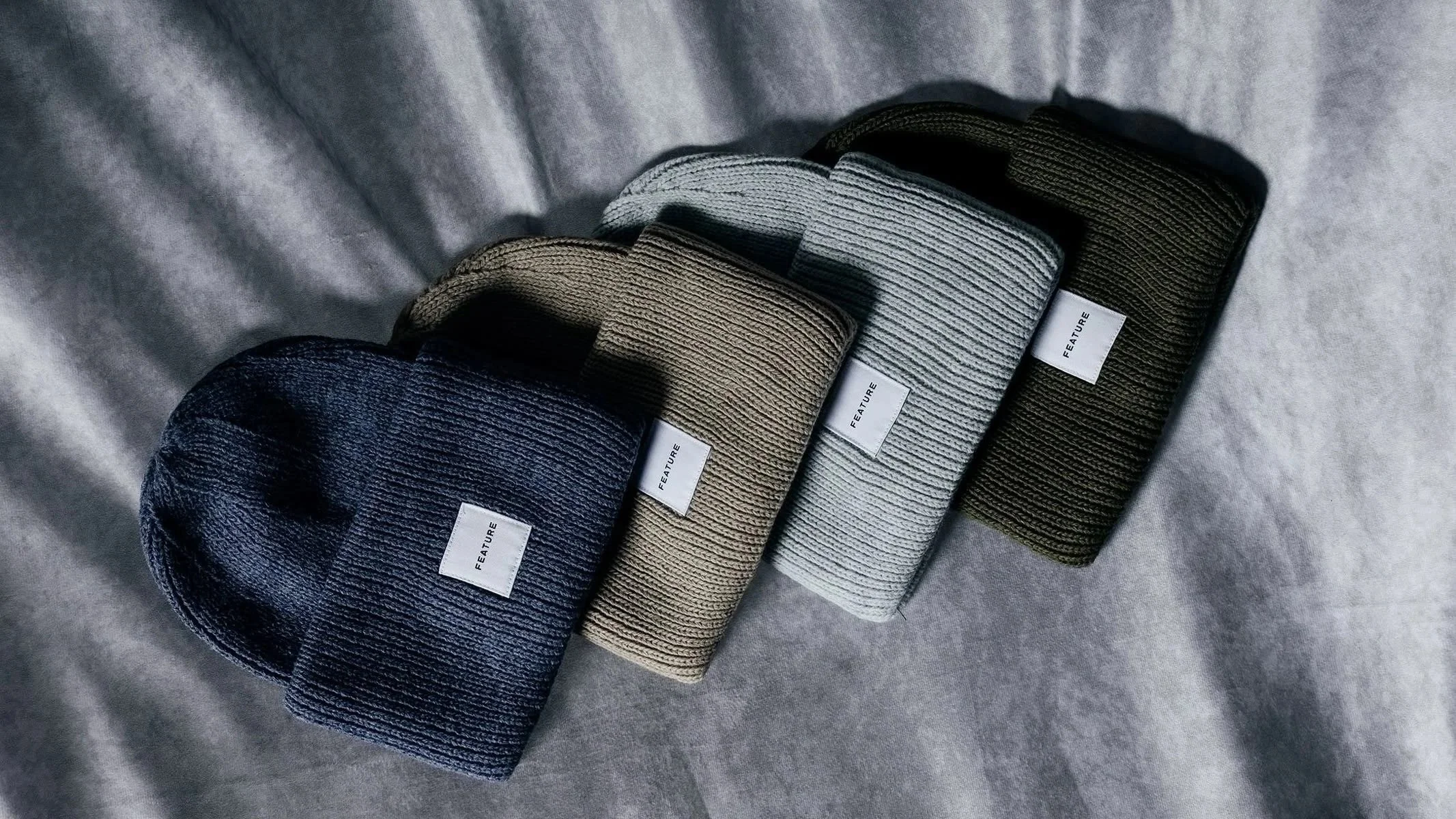 Four rolled knit beanies in various colors (navy blue, brown, gray, black) with white tags that say 'Feature', arranged on a gray fabric surface.