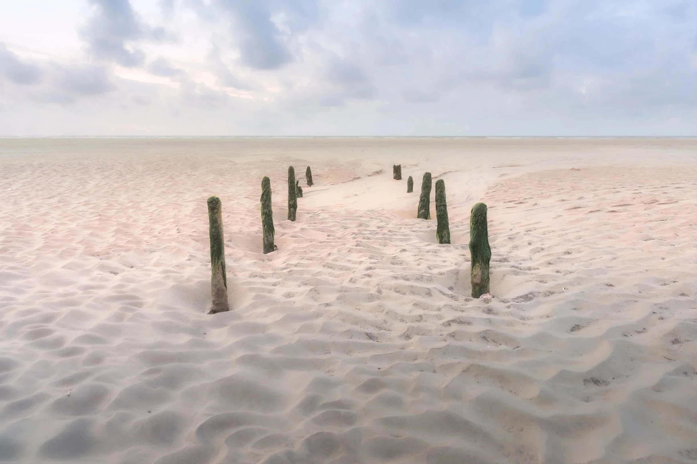 A sandy beach with wooden posts lined up in a curve leading into the distance under a cloudy sky.