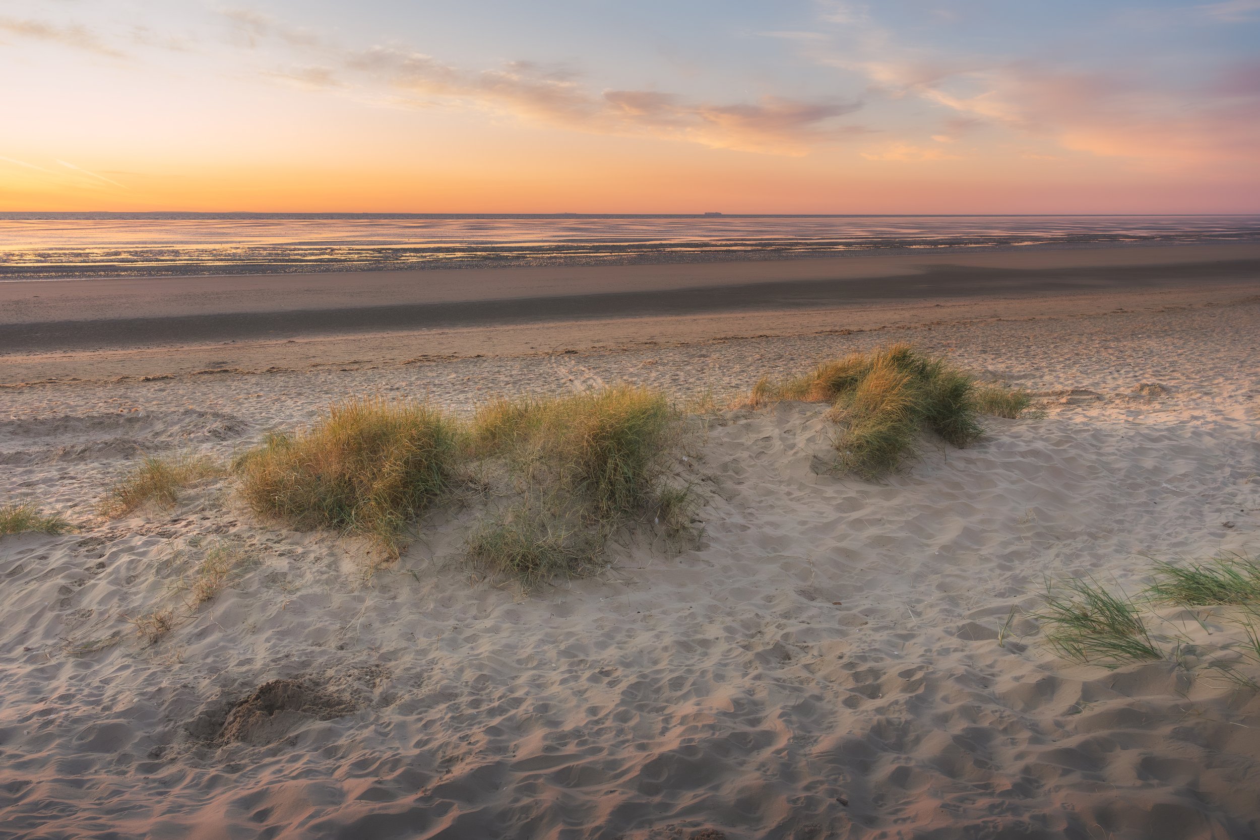 Beach scene at sunset with sand dunes, beach grass, and calm ocean waves.