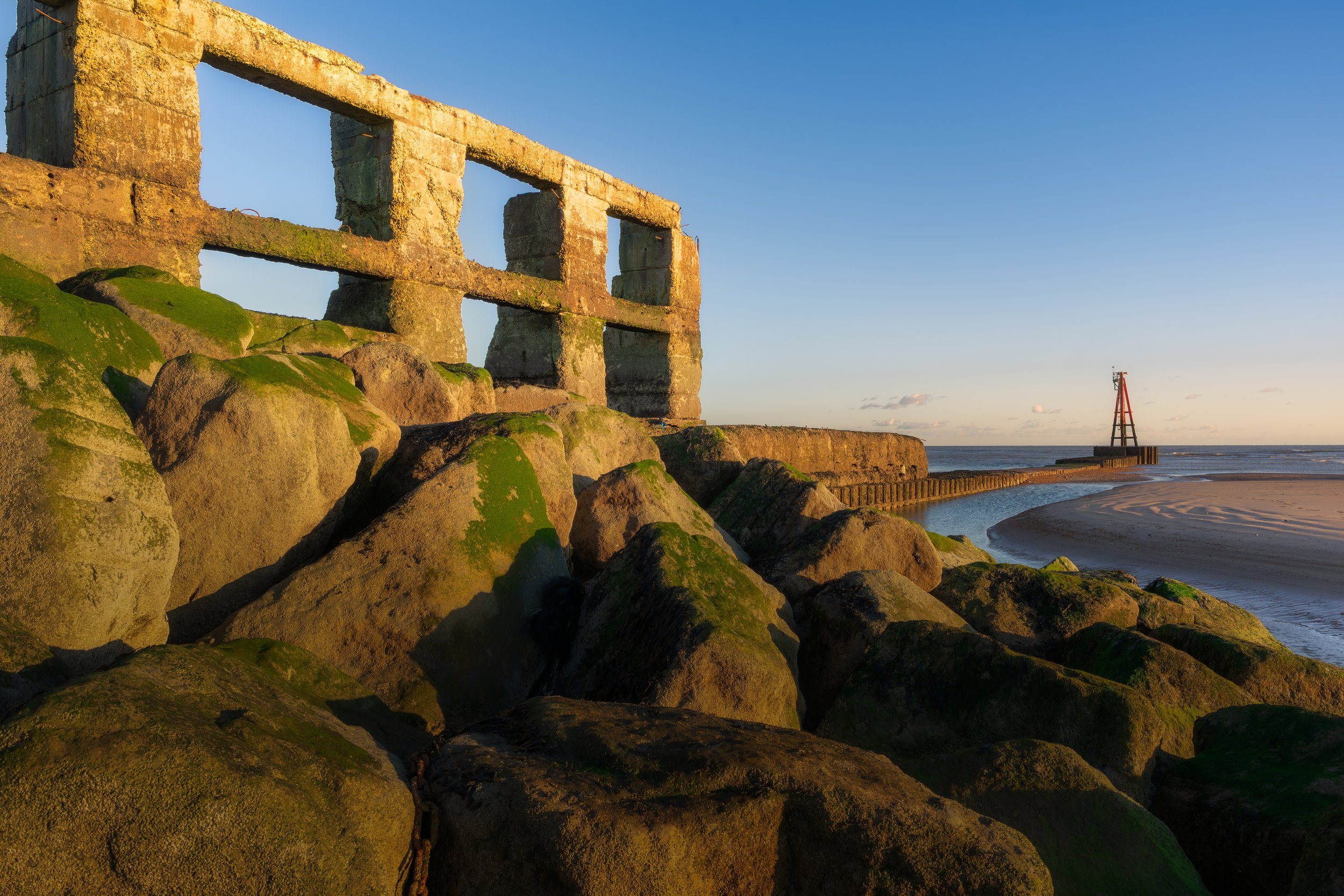 Rocky shoreline with moss-covered stones and a concrete breakwater structure, a small water channel, and a lighthouse in the distance at sunset.