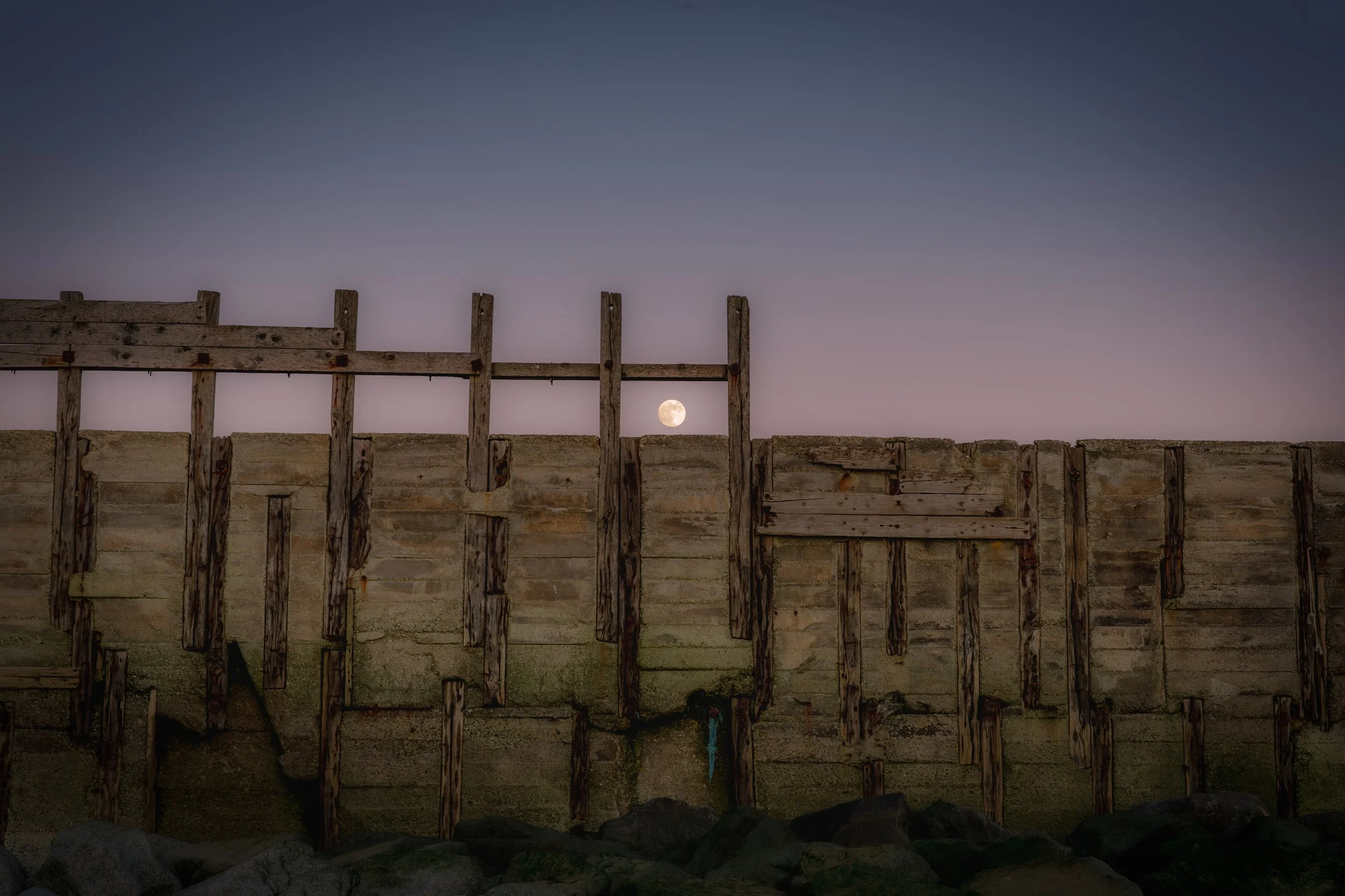 A wooden seawall or barrier with a full moon visible in the background sky at dusk or dawn.