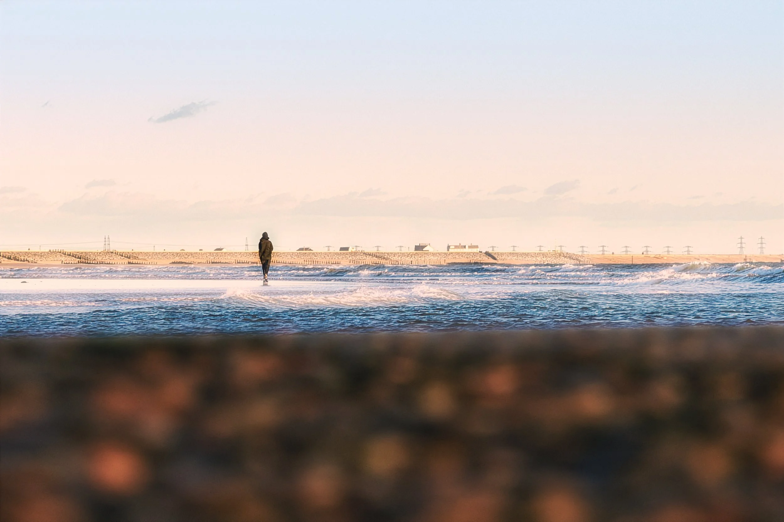A person walking along the shoreline of a beach with waves gently crashing, under a pale sky.