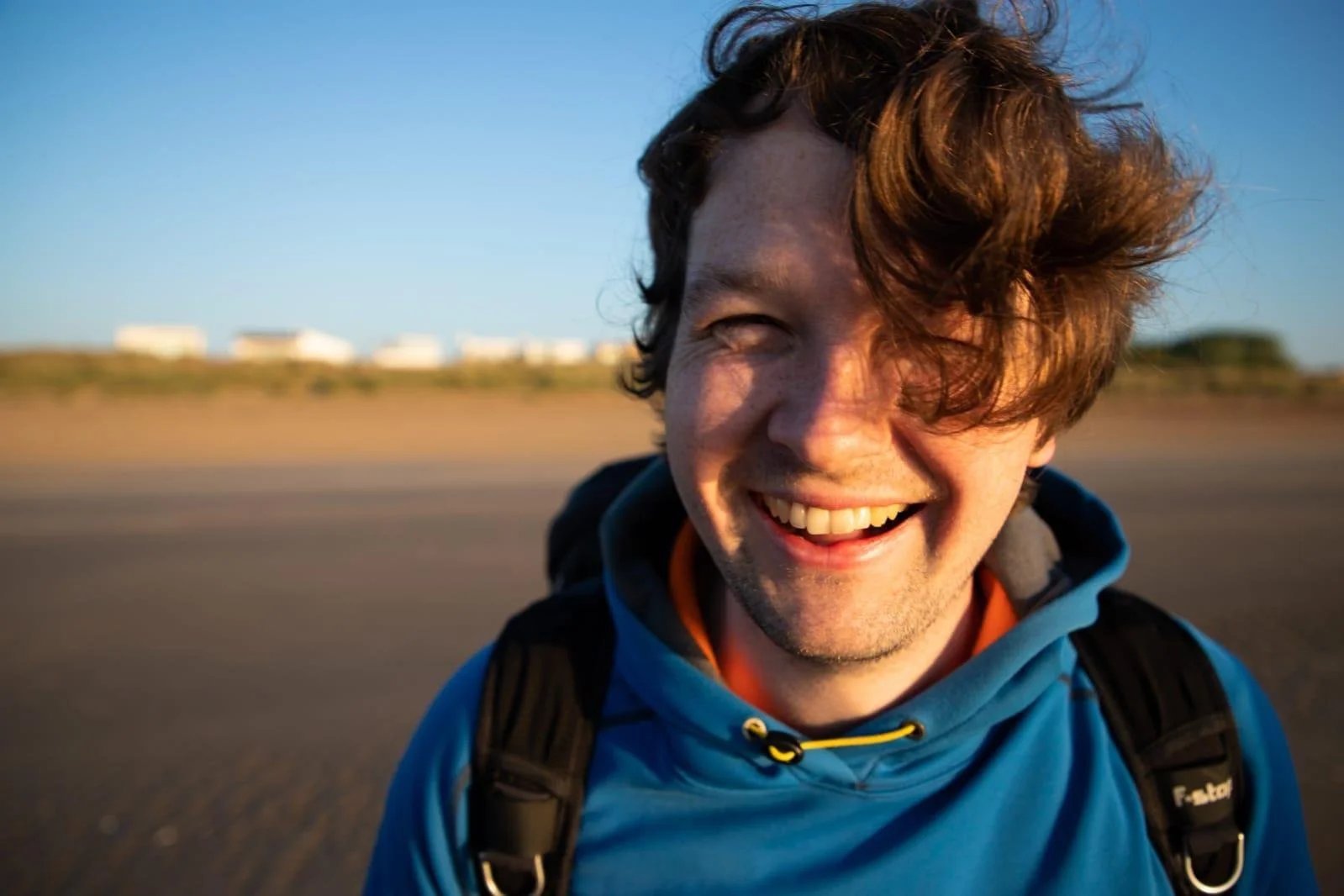 Portrait of a smiling man outdoors on a sunny day with blue skies, wearing a blue jacket and a backpack.