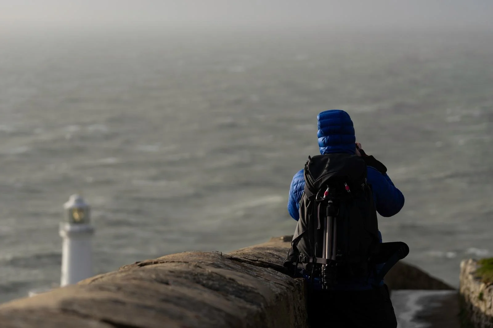 Person in blue jacket and hat sitting on a stone wall, facing the ocean, taking a photo or looking at a lighthouse in the distance.