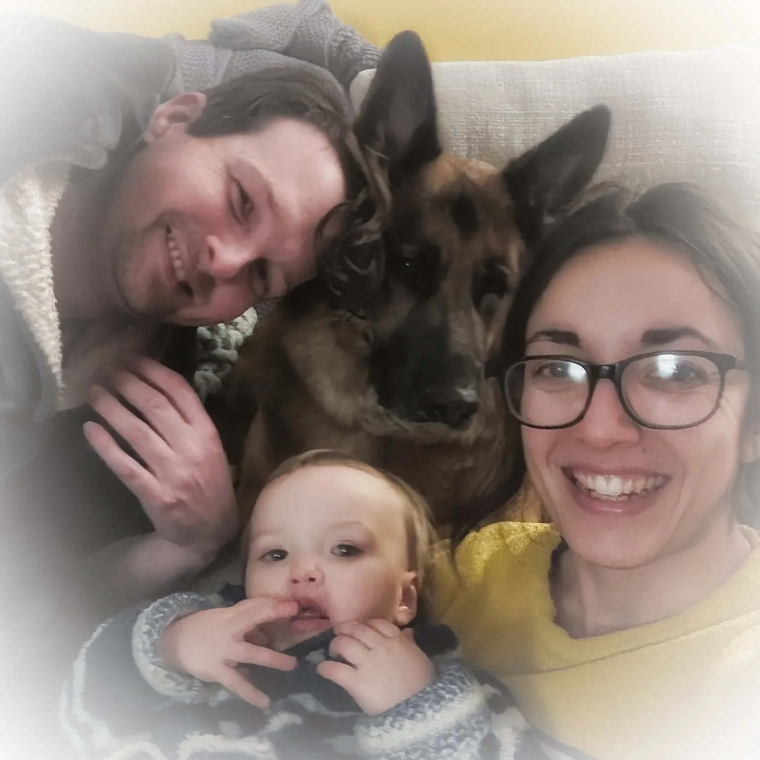 Four people, two adults and two children, lying close together with a dog, smiling and looking at the camera.