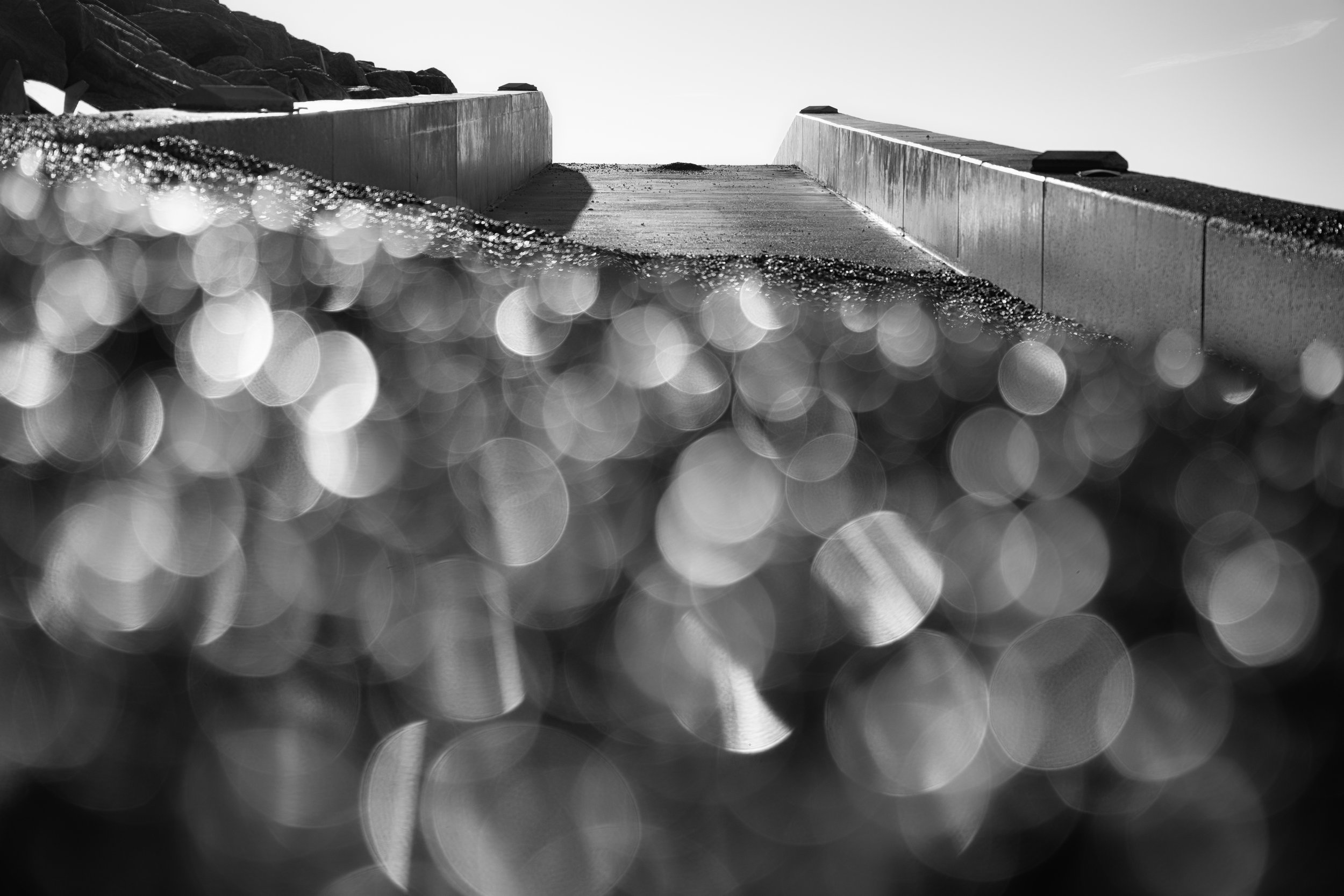 Black and white photograph of a concrete ramp or bridge viewed from below, with the sky in the background. The image features blurred bokeh effects in the foreground.