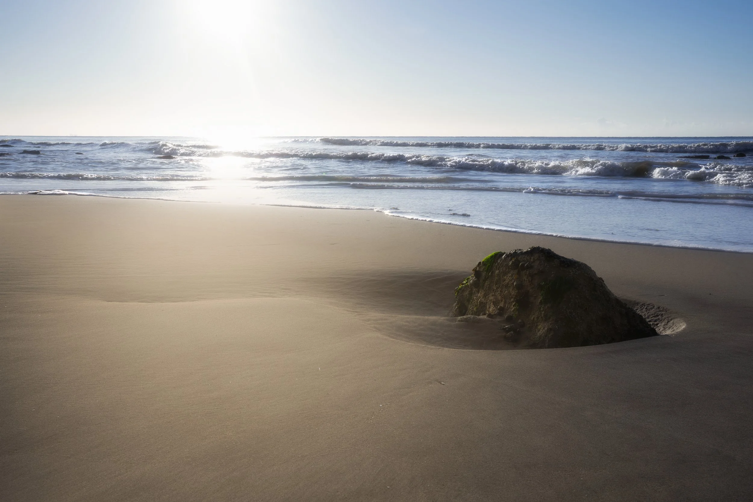 Beach with sand, a large rock covered in moss, waves in the ocean, and the sun shining in the sky.