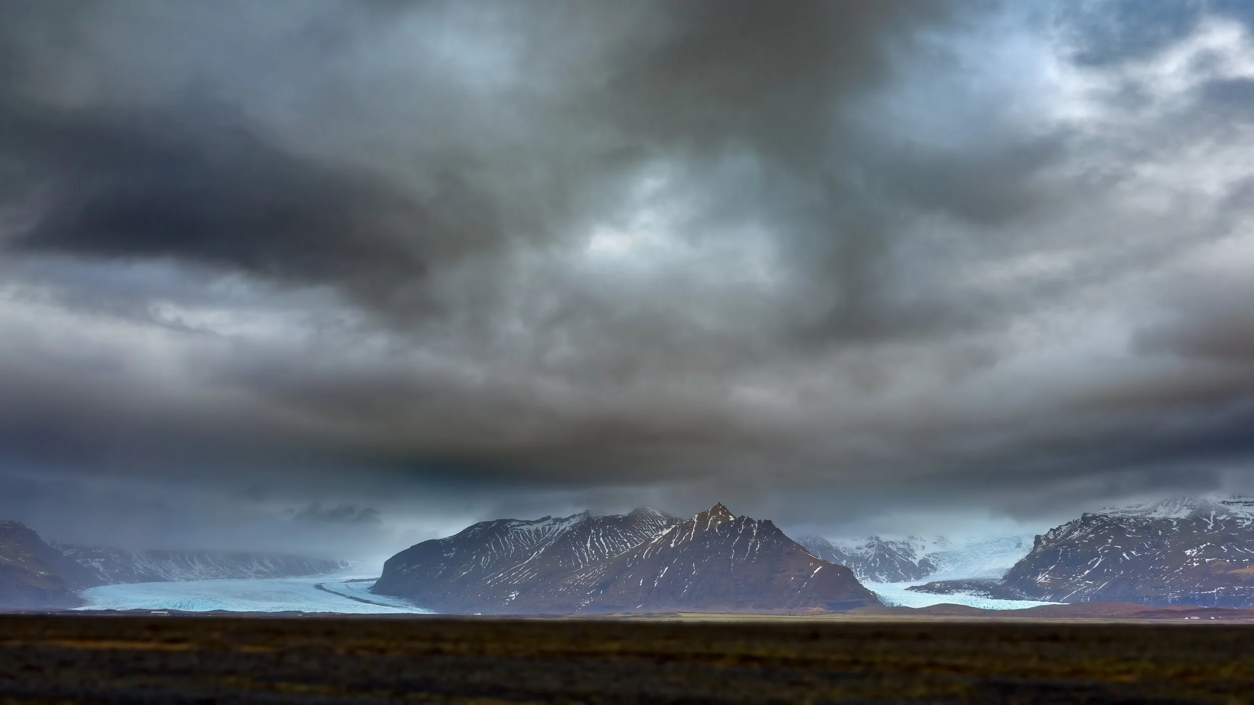 A landscape of mountains with snow patches, dark and cloudy skies, and a glacier in the distance under an overcast sky.