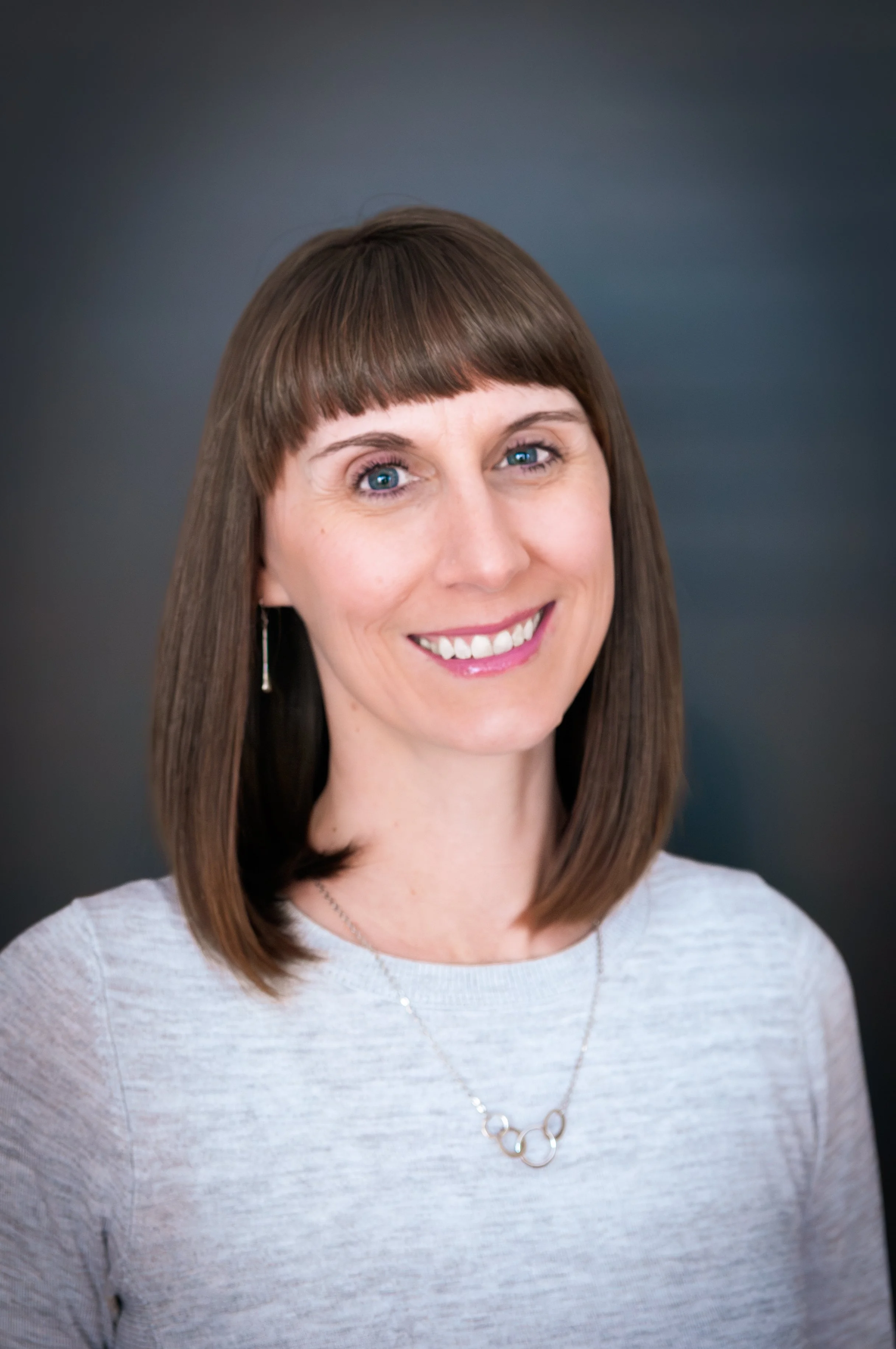 A woman with shoulder-length brown hair, blue eyes, and fair skin, smiling, wearing a light gray top, earrings, and a necklace, against a dark background.