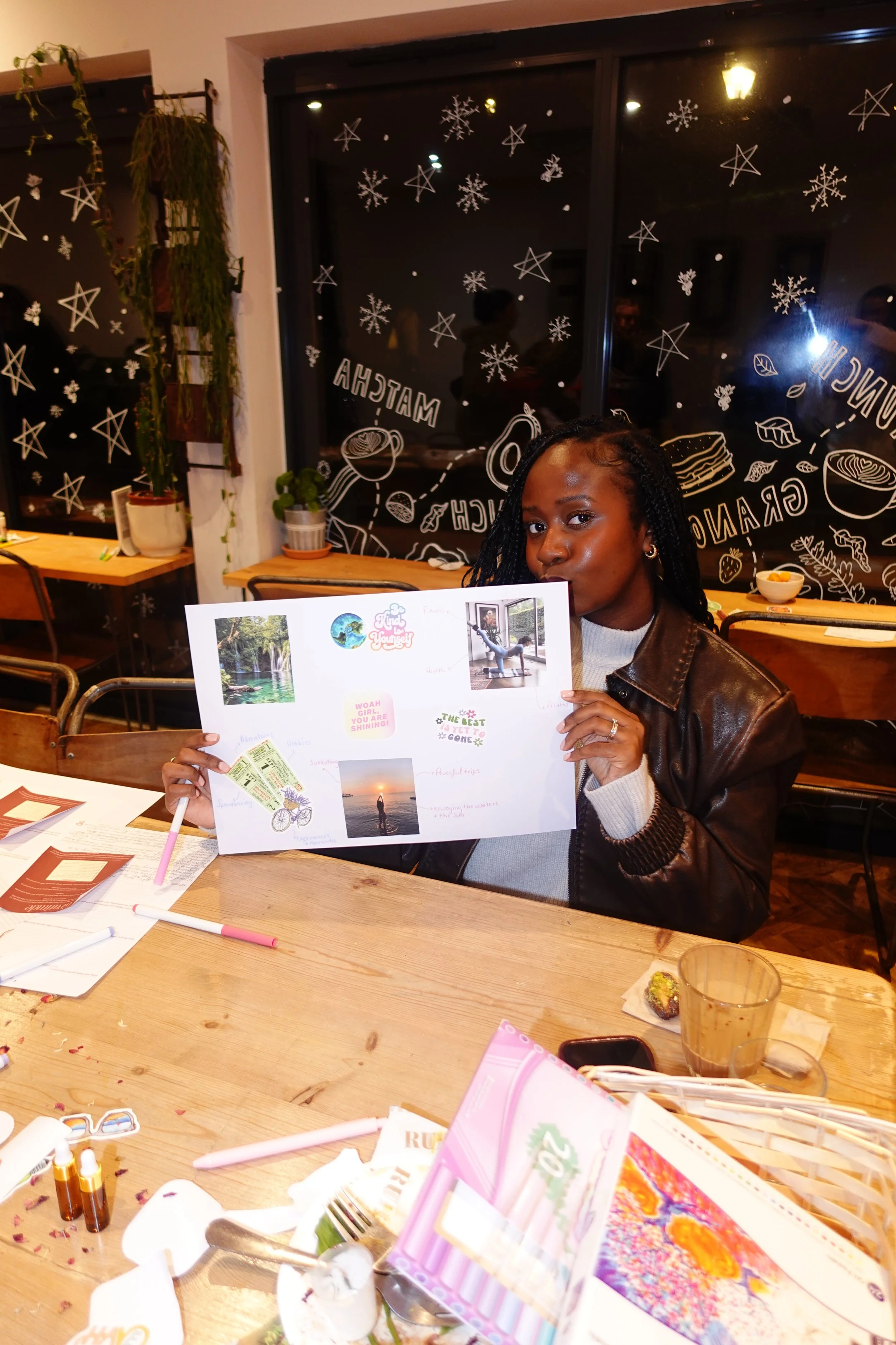 A woman sitting at a table in a cafe holding a decorated poster board with various pictures and notes, with a glittery cup and papers scattered on the table. Snowflake and holiday-themed window decorations are visible in the background.
