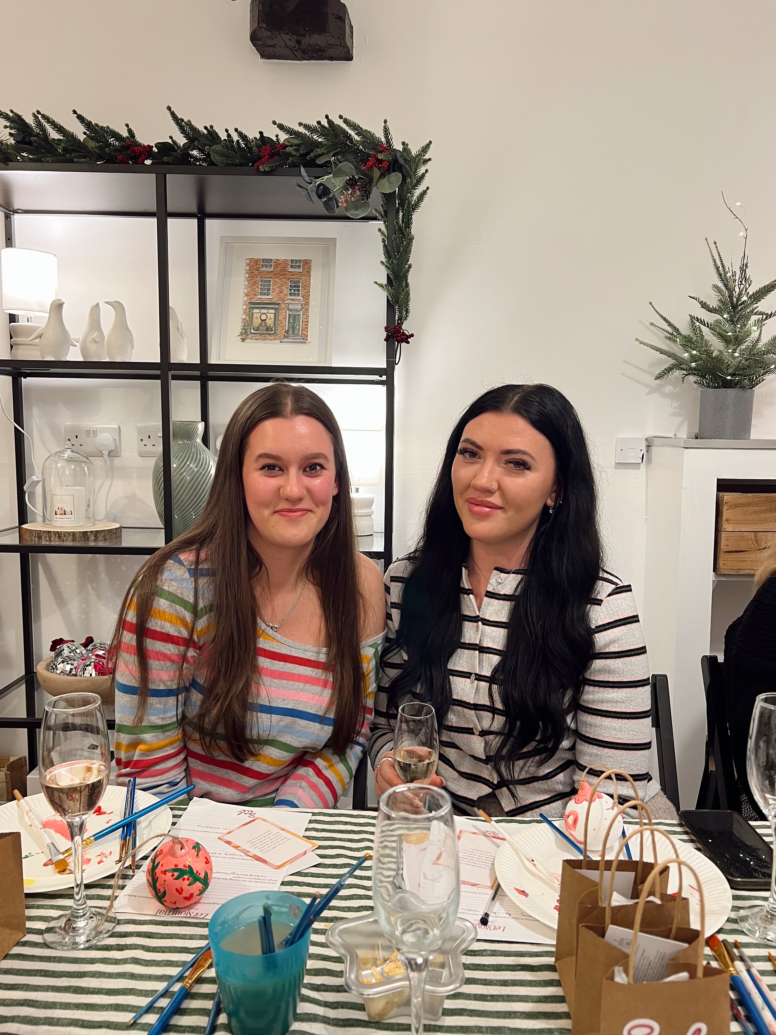 Two women sitting at a holiday-themed dinner table with decorations, drinks, and gifts, smiling for the camera, with a Christmas garland, small Christmas tree, and holiday decor in the background.