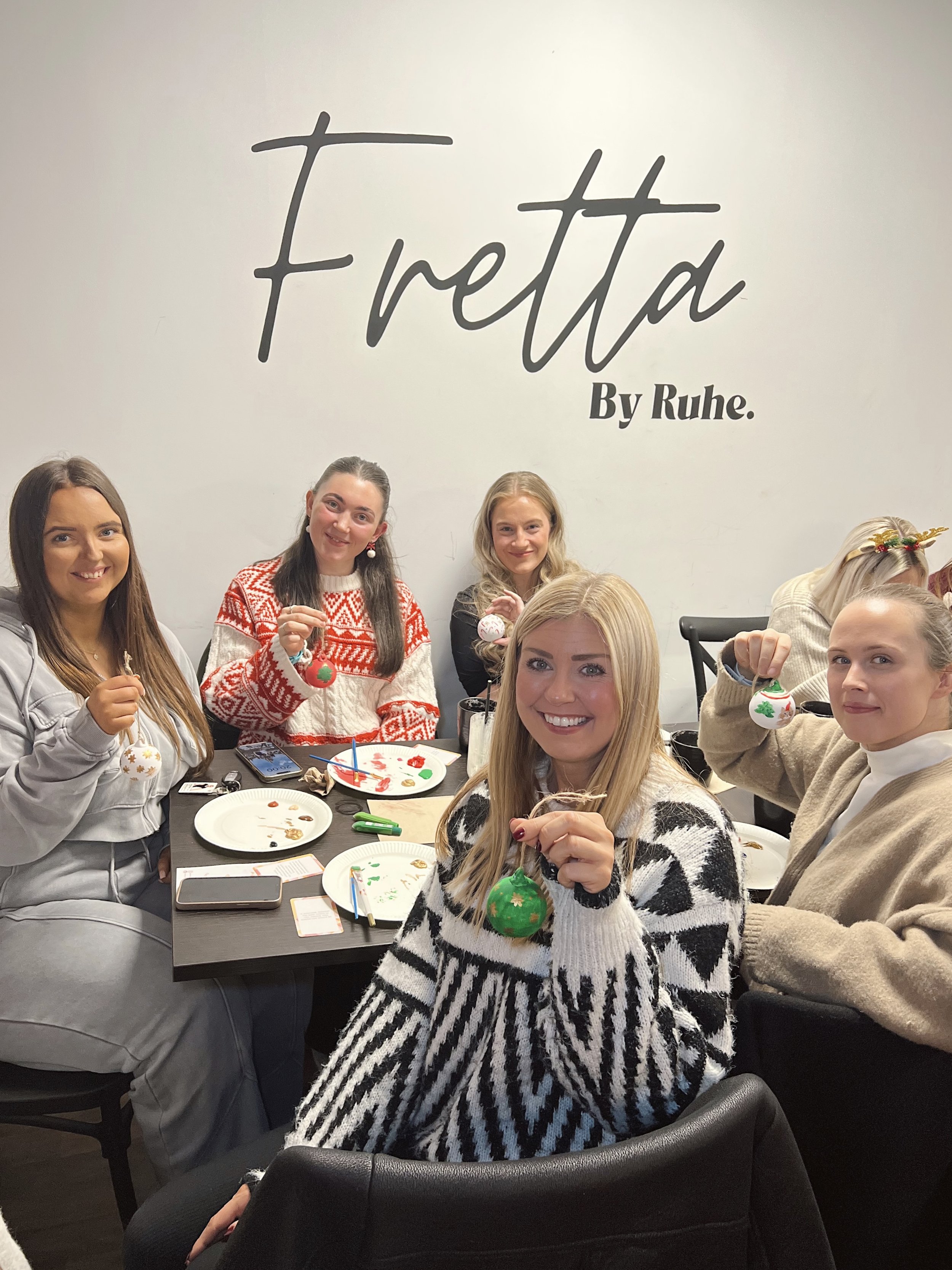 Group of women sitting at a table, holding Christmas ornaments and smiling, with Christmas decoration supplies on the table.