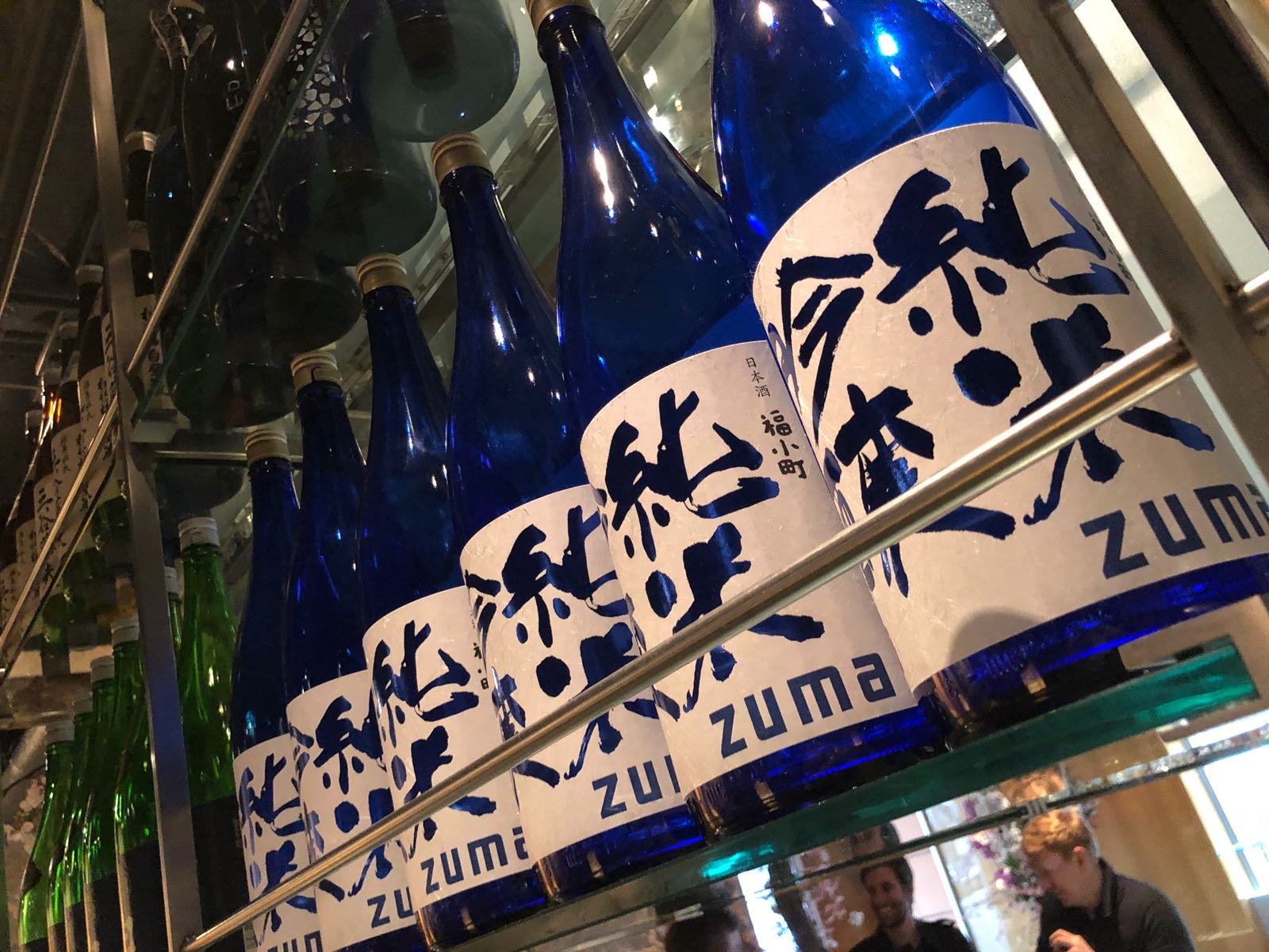 Blue bottles of sake with white labels featuring Japanese characters, on a glass shelf in a restaurant or bar.