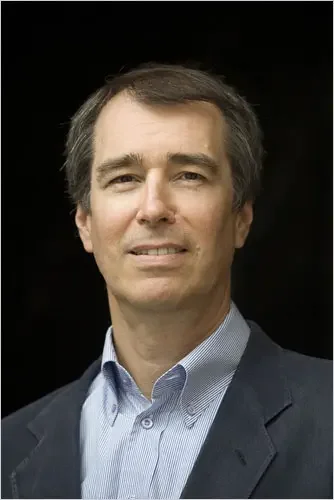 A man in a blue shirt and dark blazer against a black background.