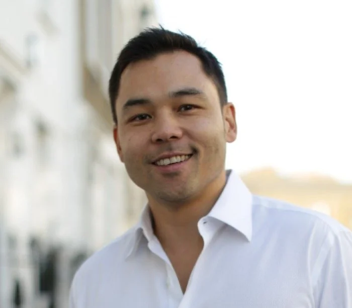 A smiling man with dark hair wearing a white button-up shirt standing outdoors in front of a blurred cityscape.