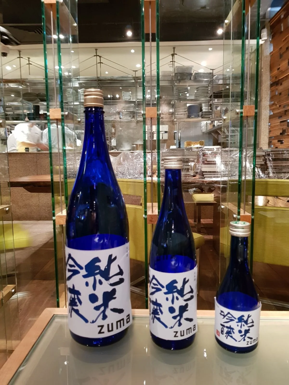 Three blue bottles with white labels and Japanese characters, labeled 'ZUMA', are placed on a glass display case in front of a restaurant kitchen with chefs and shelves of plates and dishes inside.