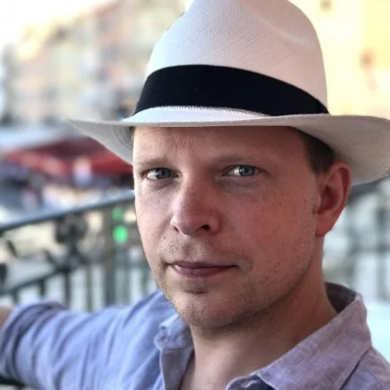 A man wearing a white fedora hat with a black band, looking at the camera, with a blurred outdoor background.