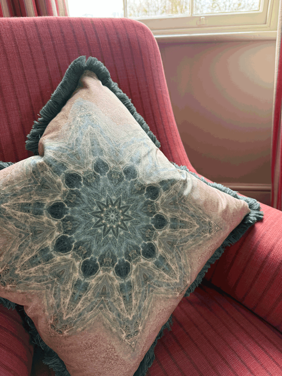 Decorative pillow with a detailed mandala-like pattern resting on a red upholstered armchair near a window.
