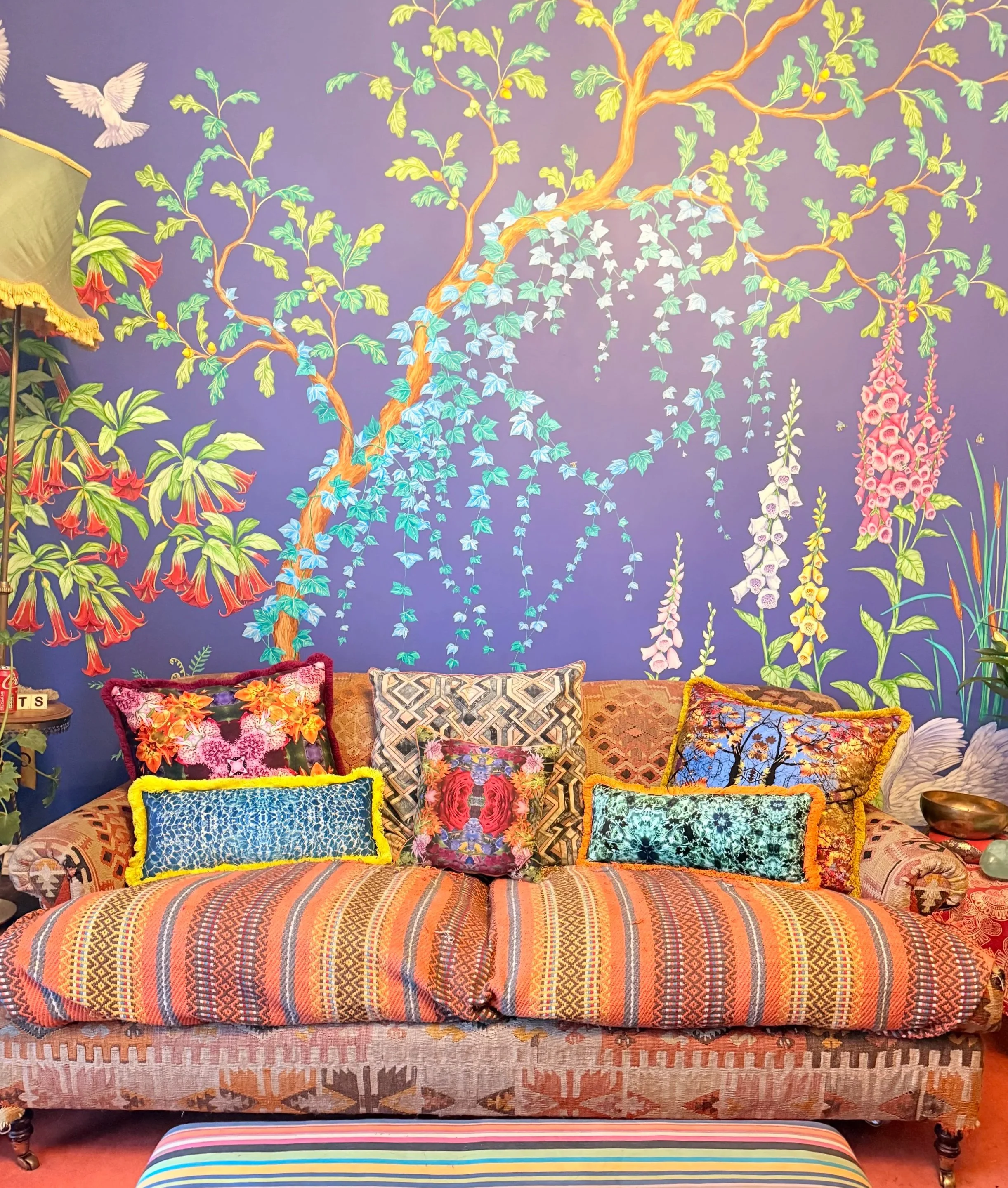 Colorful living room sofa with multiple patterned and floral cushions against a vibrant mural of flowering plants and greenery on the wall.