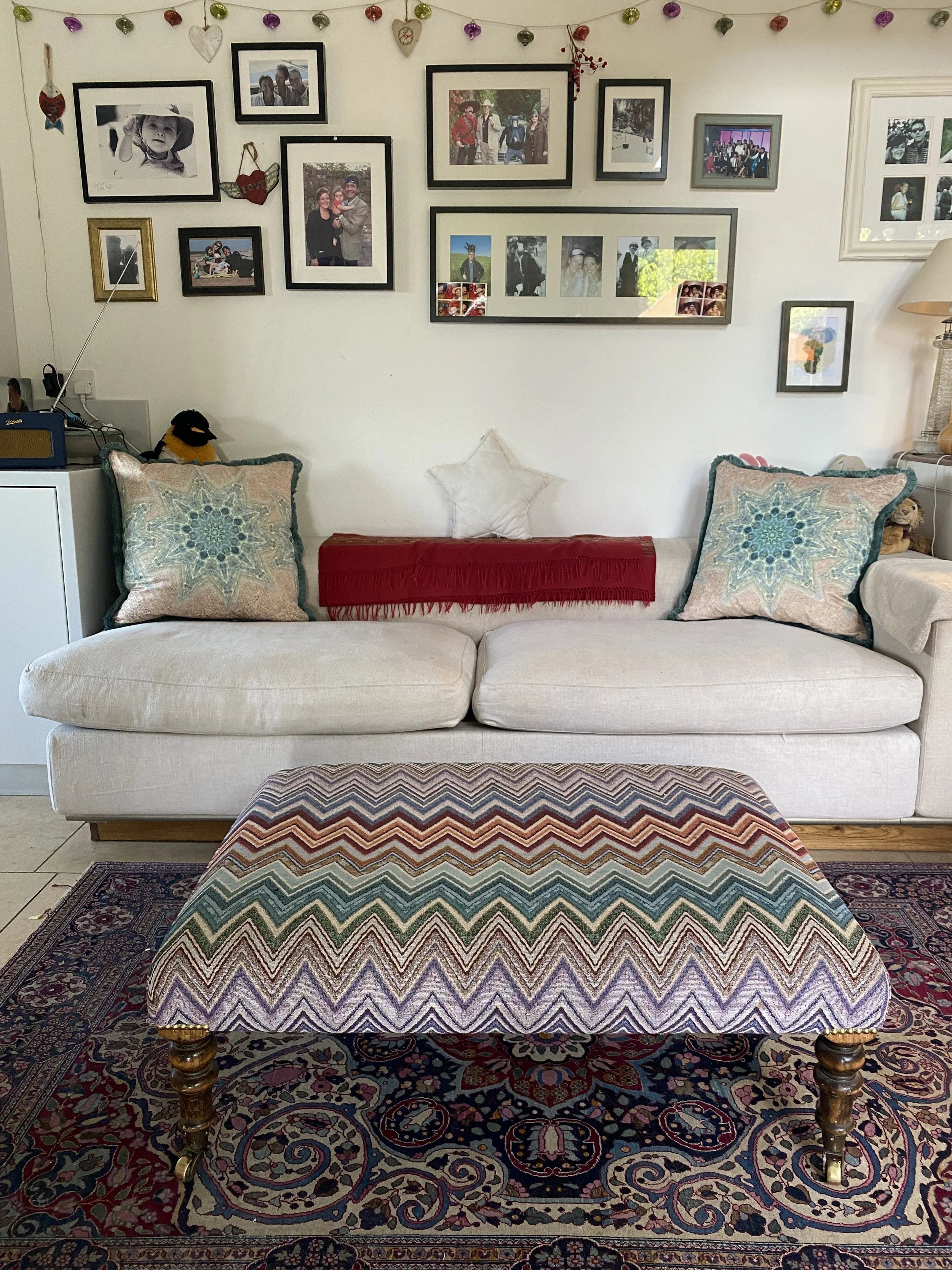 A living room with a white sofa, decorated with two patterned pillows, a red scarf, and a star-shaped pillow. In front of the sofa, there is a colorful patterned ottoman with wooden legs on a Persian-style rug. The wall behind the sofa displays an array of framed photographs and artwork, with garlands of hanging hearts and ornaments. To the left, there's a small white cabinet with a yellow speaker and a stuffed penguin, and to the right, a lamp on a side table.