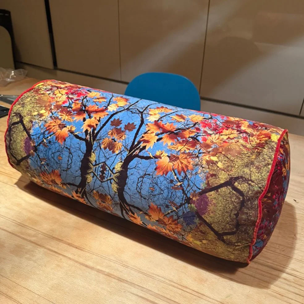 A cylindrical decorative pillow with a tree and autumn leaves pattern, placed on a wooden table with a blue chair and beige cabinets in the background.
