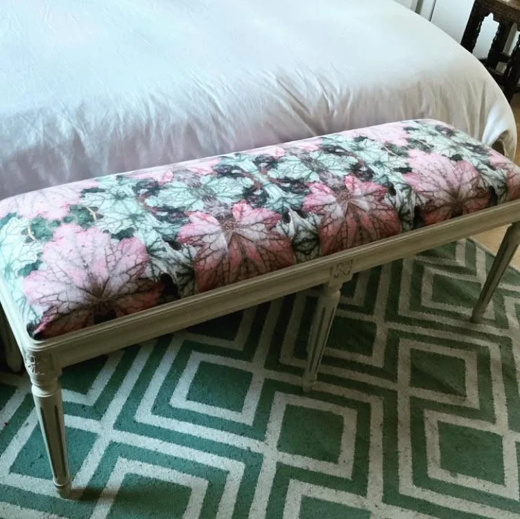A bench with a cushion covered in a floral and foliage pattern, placed at the foot of a bed with white bedding, on a geometric-patterned green and white rug.