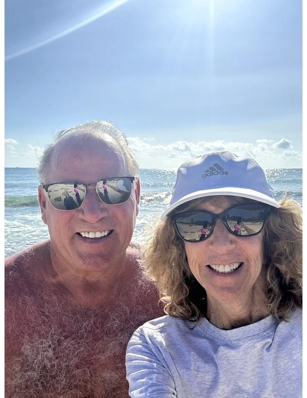 A smiling older man and woman taking a selfie on a beach, both wearing sunglasses. The man is shirtless with white hair, and the woman is wearing a light gray shirt, a white cap, and sunglasses. The ocean and blue sky are visible in the background.