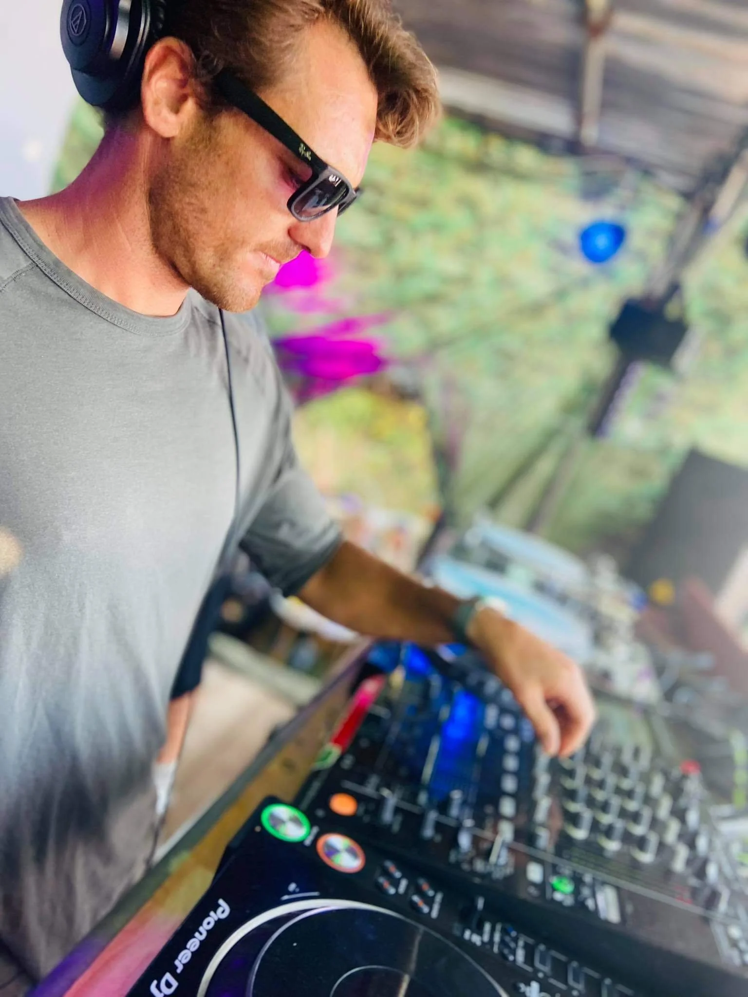 A male DJ wearing sunglasses and headphones, focusing on mixing music at an outdoor event with colorful lights and trees in the background.