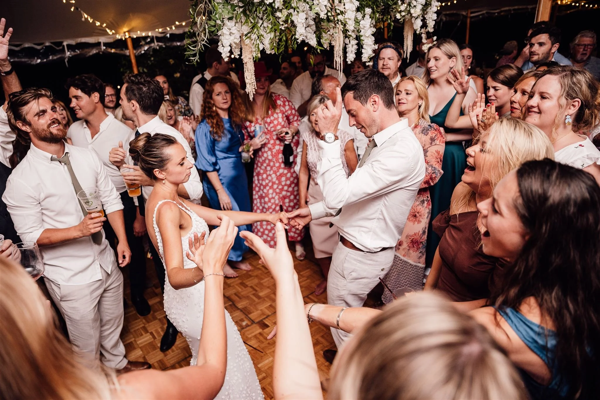 People dancing and celebrating at a wedding reception, with a bride and groom in the center holding hands while enjoying the moment, surrounded by friends and family under a decorated tent.