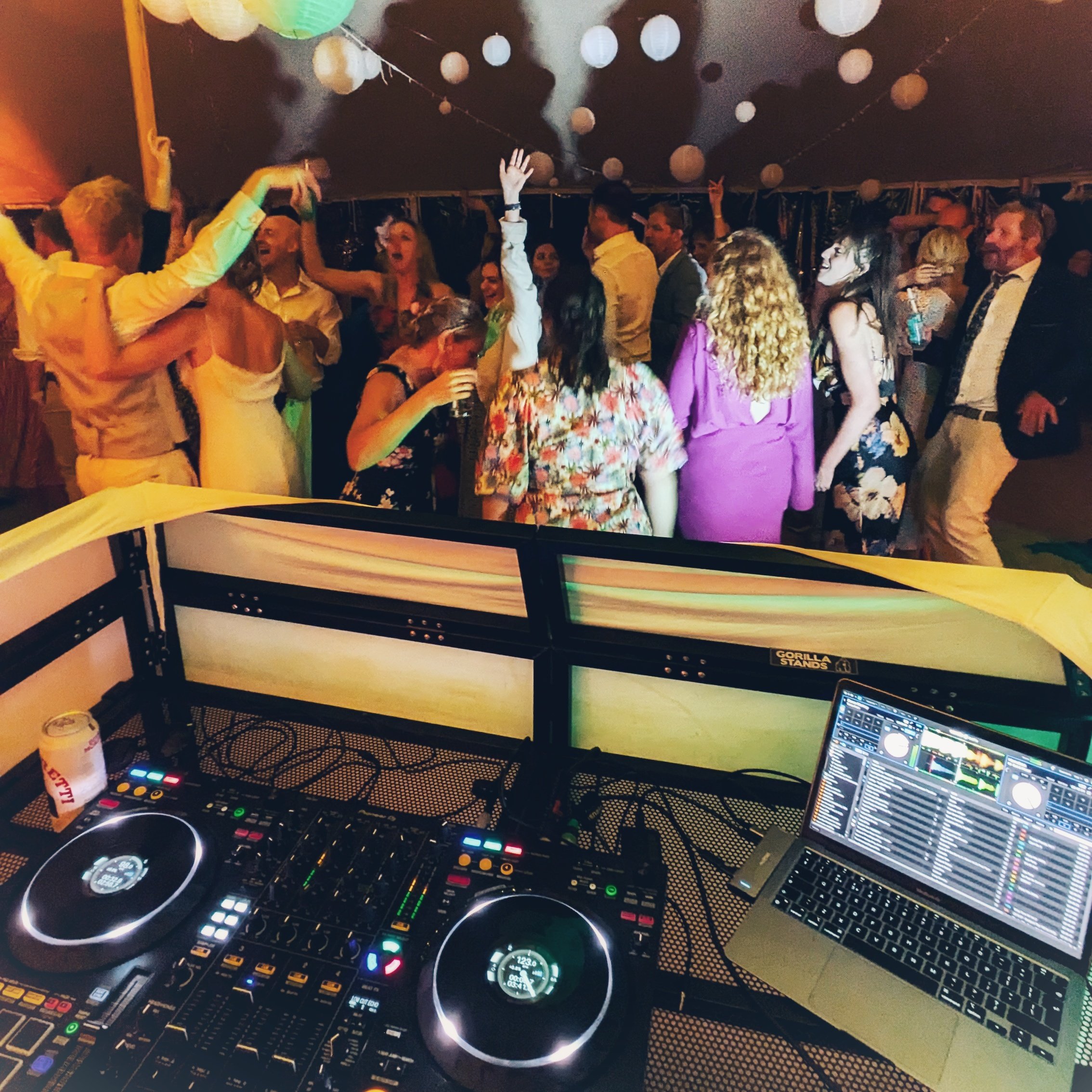 A DJ setup with turntables, a laptop, and a mixer overlooking a dance floor filled with people dancing and enjoying a party or celebration event under a tent decorated with paper lanterns.
