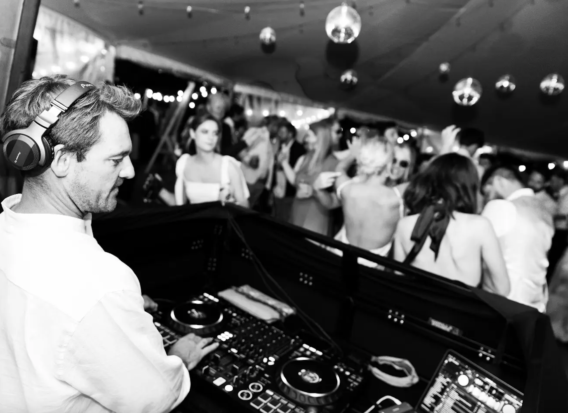 A DJ wearing headphones operates a mixing console at a party or nightclub. In the background, people dance and socialize under hanging disco balls and string lights.