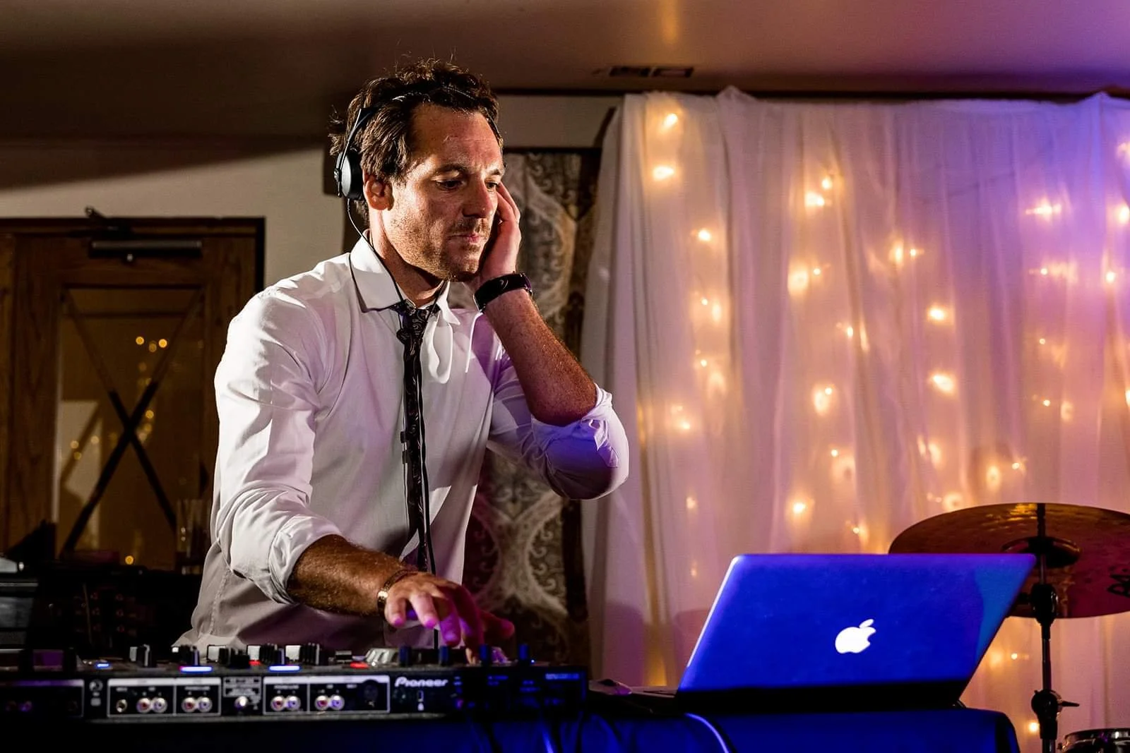 A man DJing at a party, wearing headphones, standing behind a DJ controller and a laptop, with decorative string lights in the background.