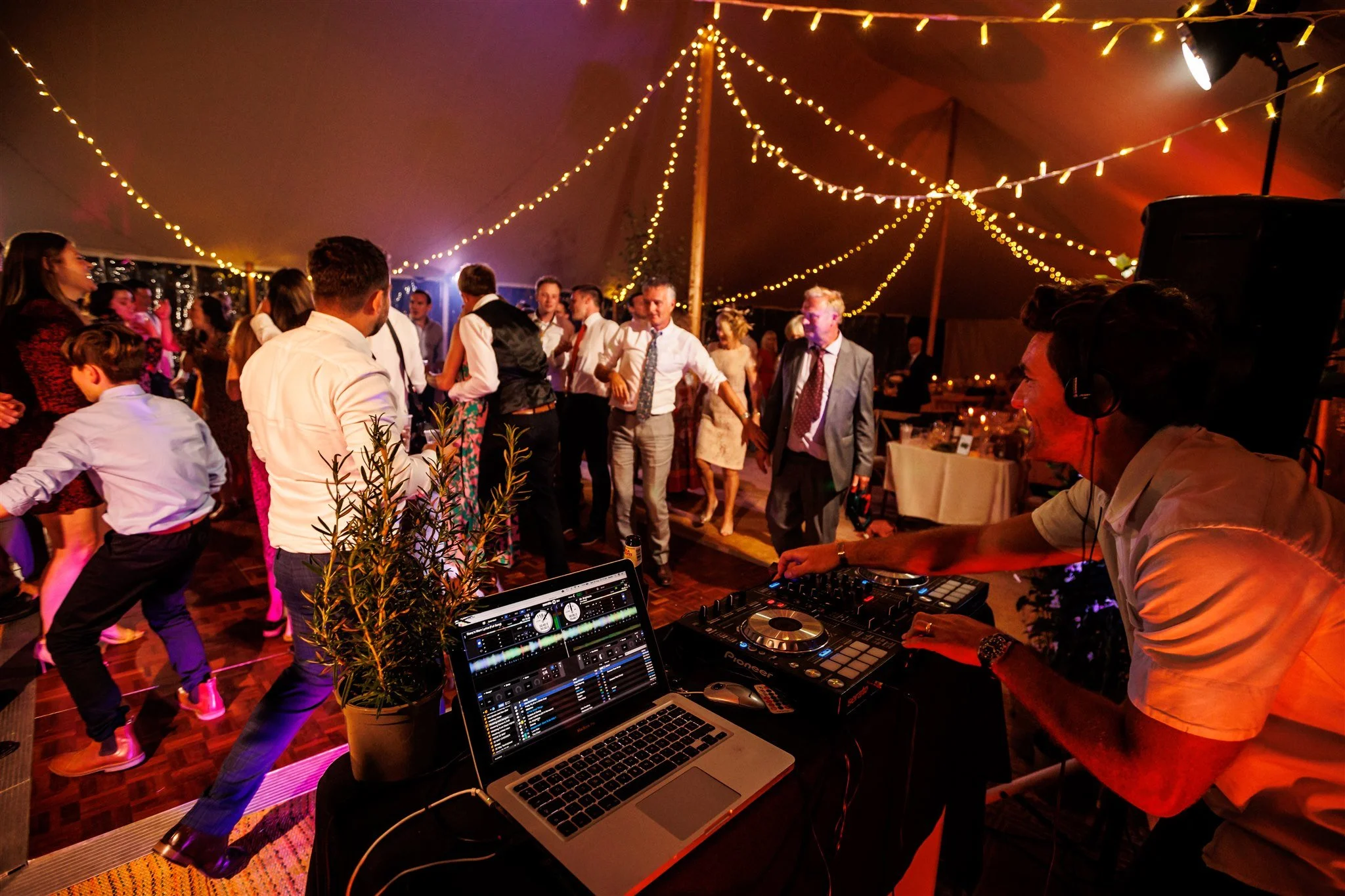 DJ in a white shirt playing music at a wedding reception with guests dancing under string lights inside a tent.