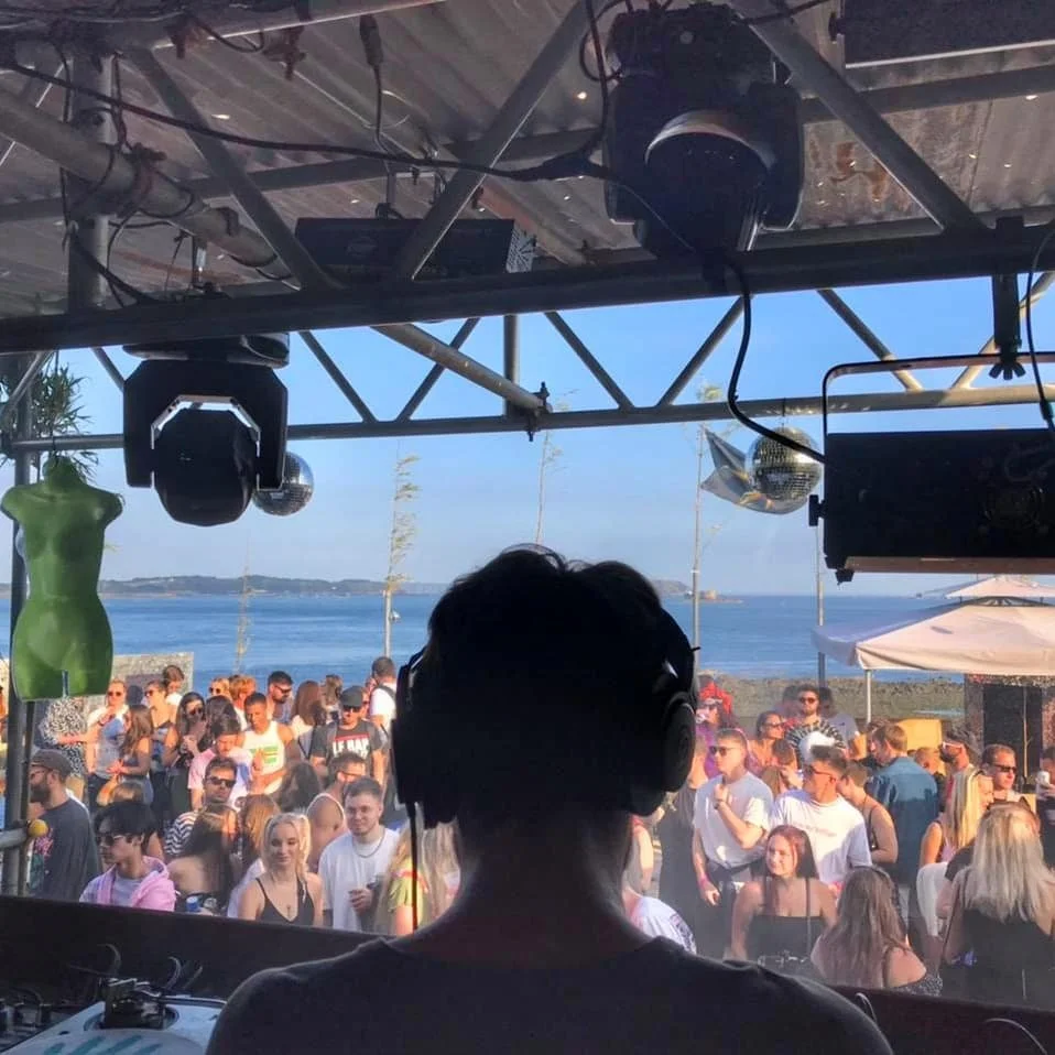 A DJ performing at a seaside outdoor event with a crowd of people dancing and socializing under a clear blue sky.