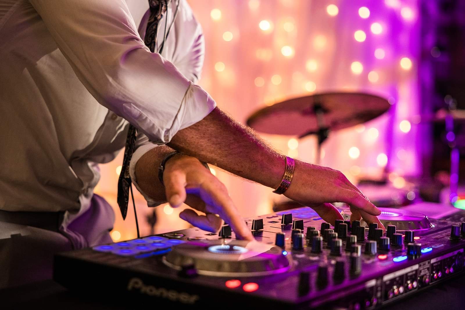 Close-up of a DJ's hands manipulating turntables and controls at a party or event with colorful lighting and blurred background.