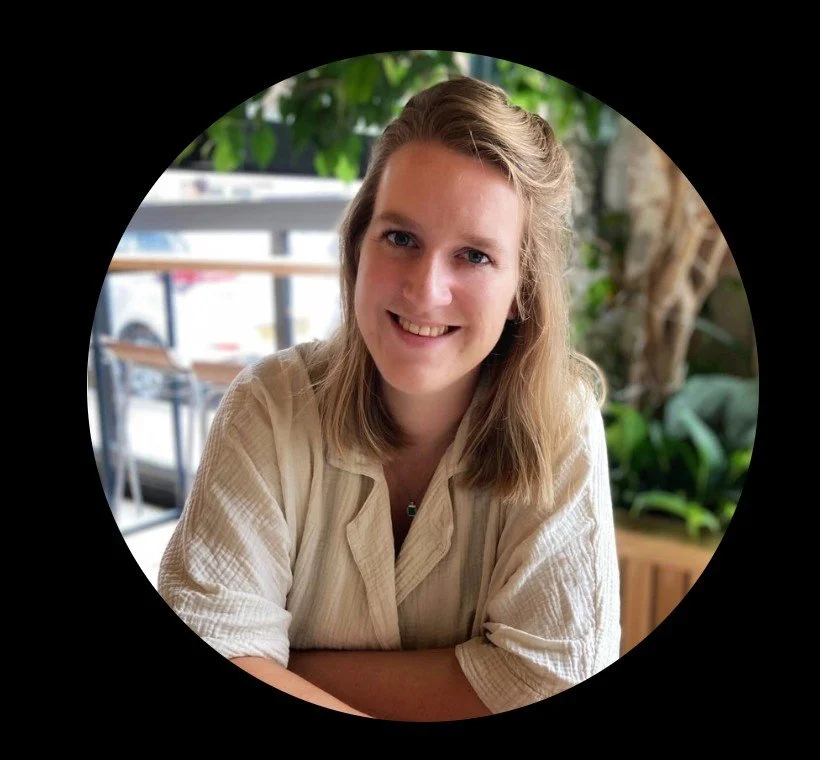 A young woman with shoulder-length blonde hair smiling at the camera, sitting at a table in a cafe or restaurant with greenery in the background.