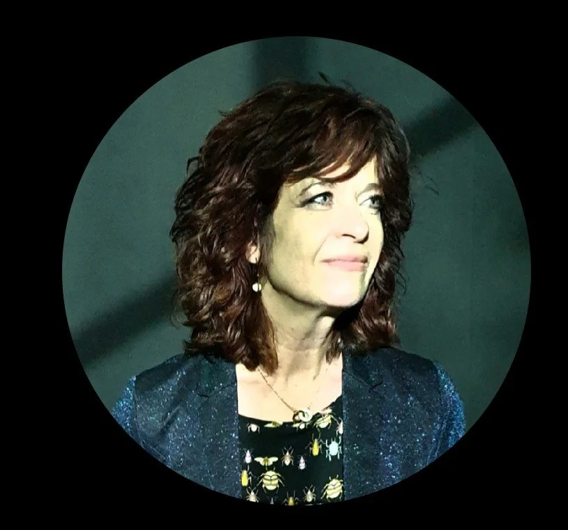 A woman with shoulder-length, curly brown hair, wearing a dark blue sparkly blazer and a black shirt with a yellow bee pattern, looking slightly to the side.