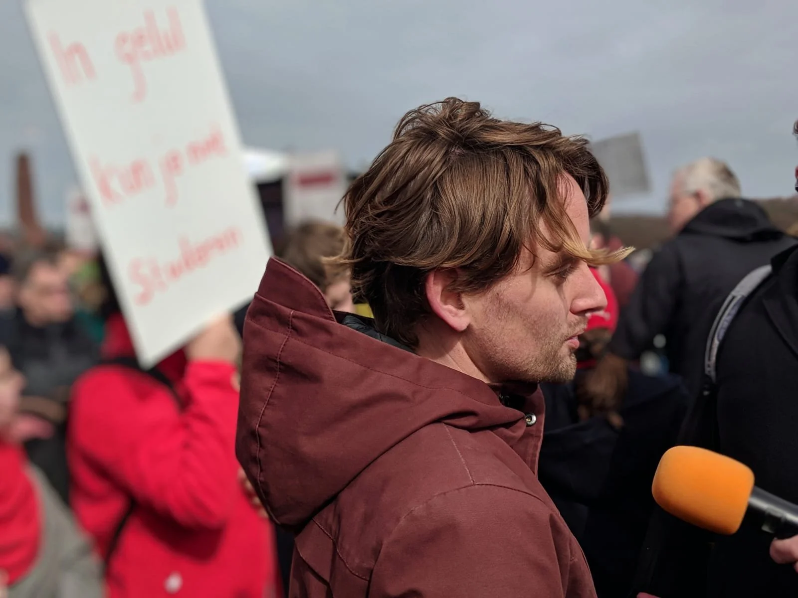 A man with brown hair and a beard standing in a crowd during an outdoor event, wearing a red jacket. People around him hold signs and appear to be protesting or gathering.