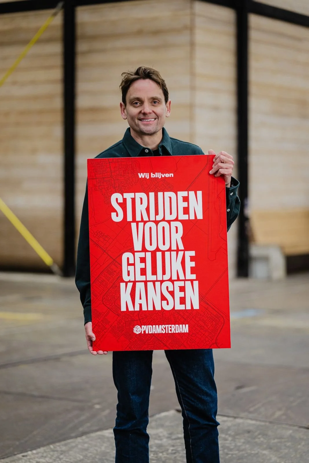 A man standing indoors in front of a wooden wall, holding a red sign that reads "Wij blijven strijden voor gelijke kansen" and "PVD Amsterdam."