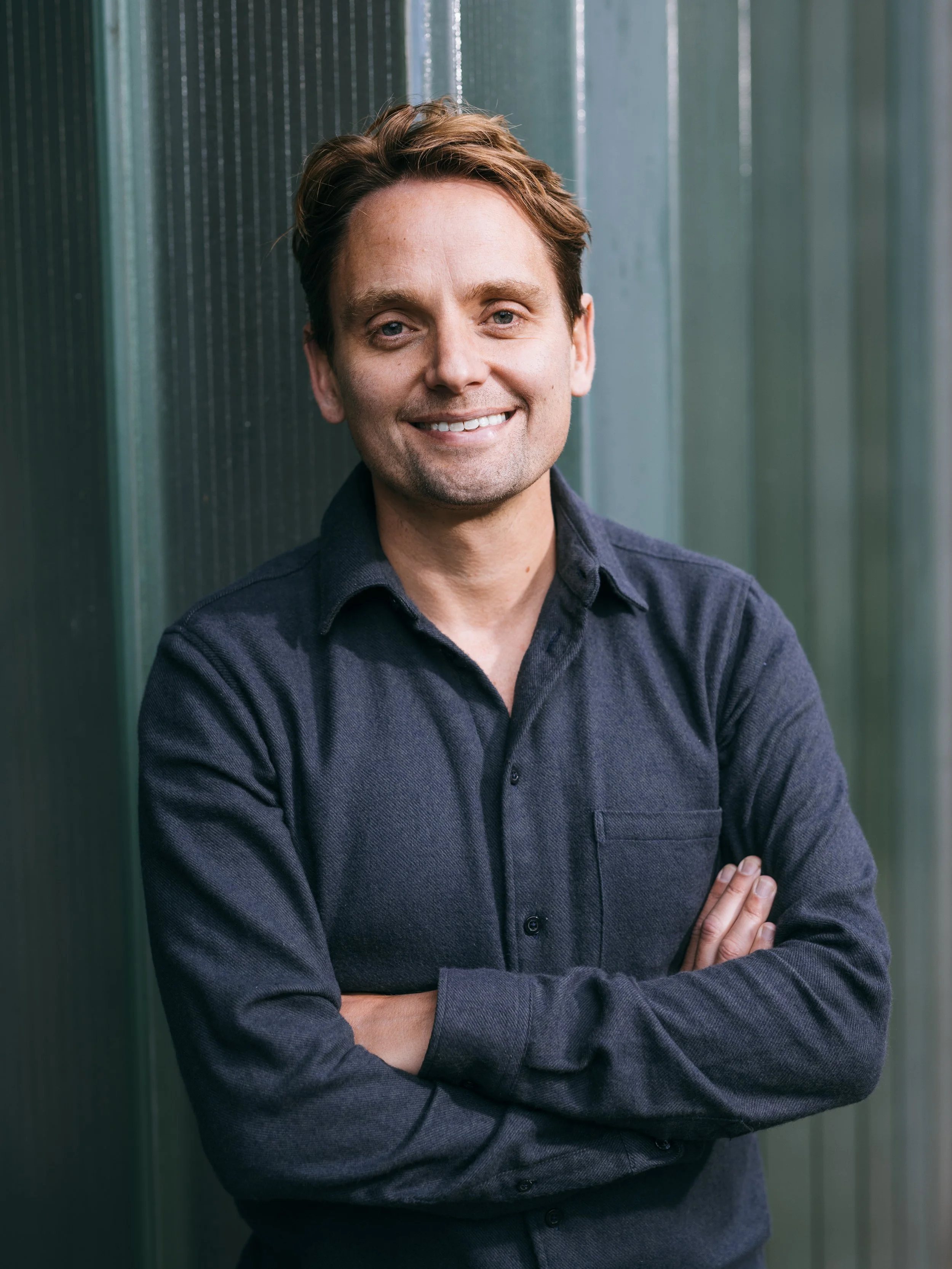 A smiling man with light skin, brown hair, dressed in a dark button-up shirt, standing with arms crossed in front of a green wooden wall.