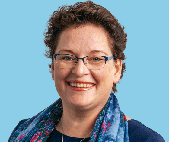 Close-up portrait of a woman with short curly hair, glasses, smiling, wearing a blue scarf and dark top, against a light blue background.