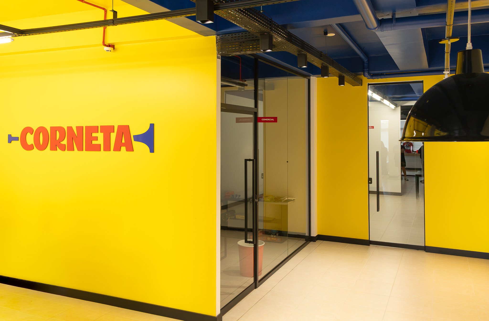 Entrada de loja com paredes coloridas amarelo e azul, com portas de vidro, sinalização de 'Comercial' na porta.