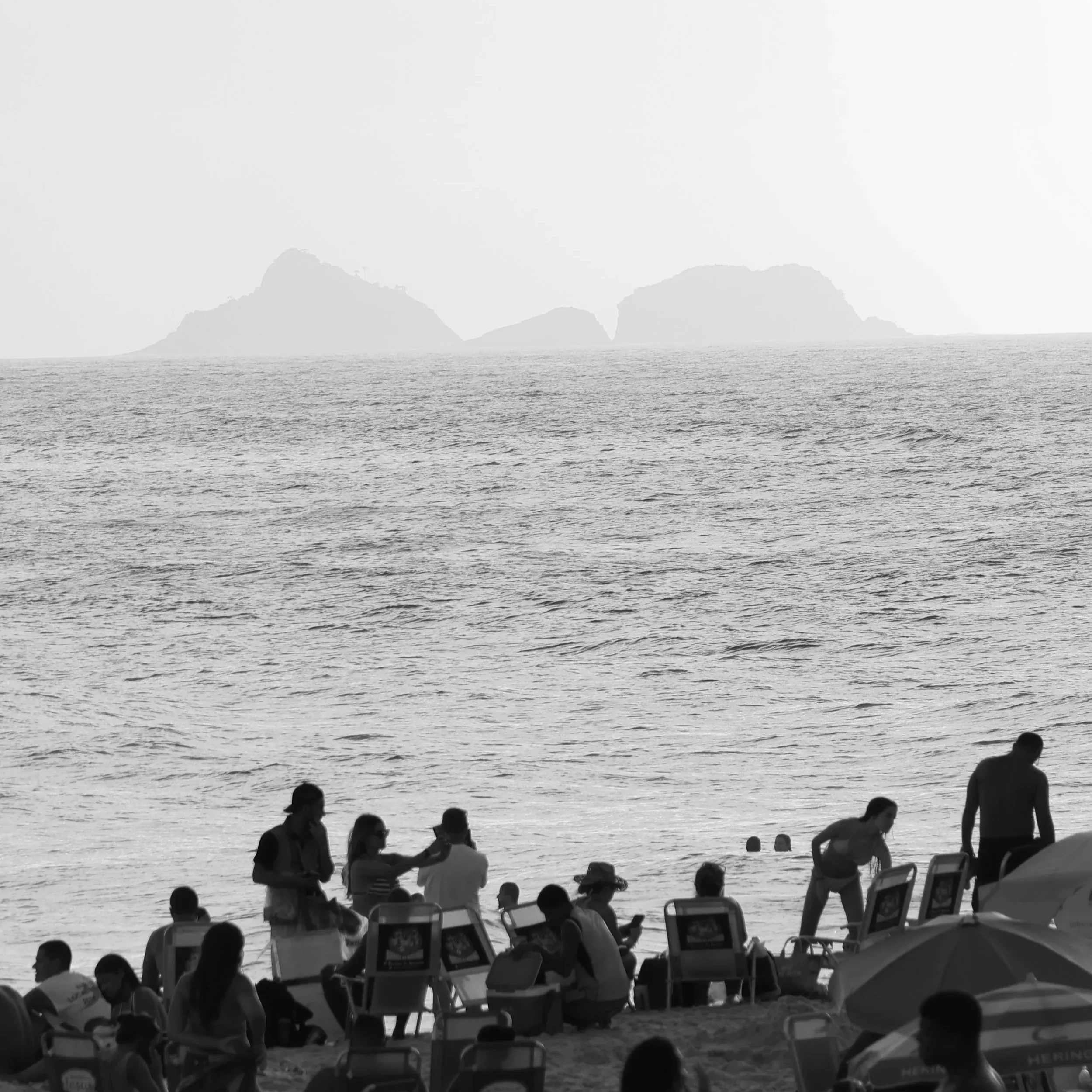 Pessoas na praia, algumas sentadas em cadeiras, outras de pé, com o mar ao fundo e morros ao longe, em uma cena em preto e branco.