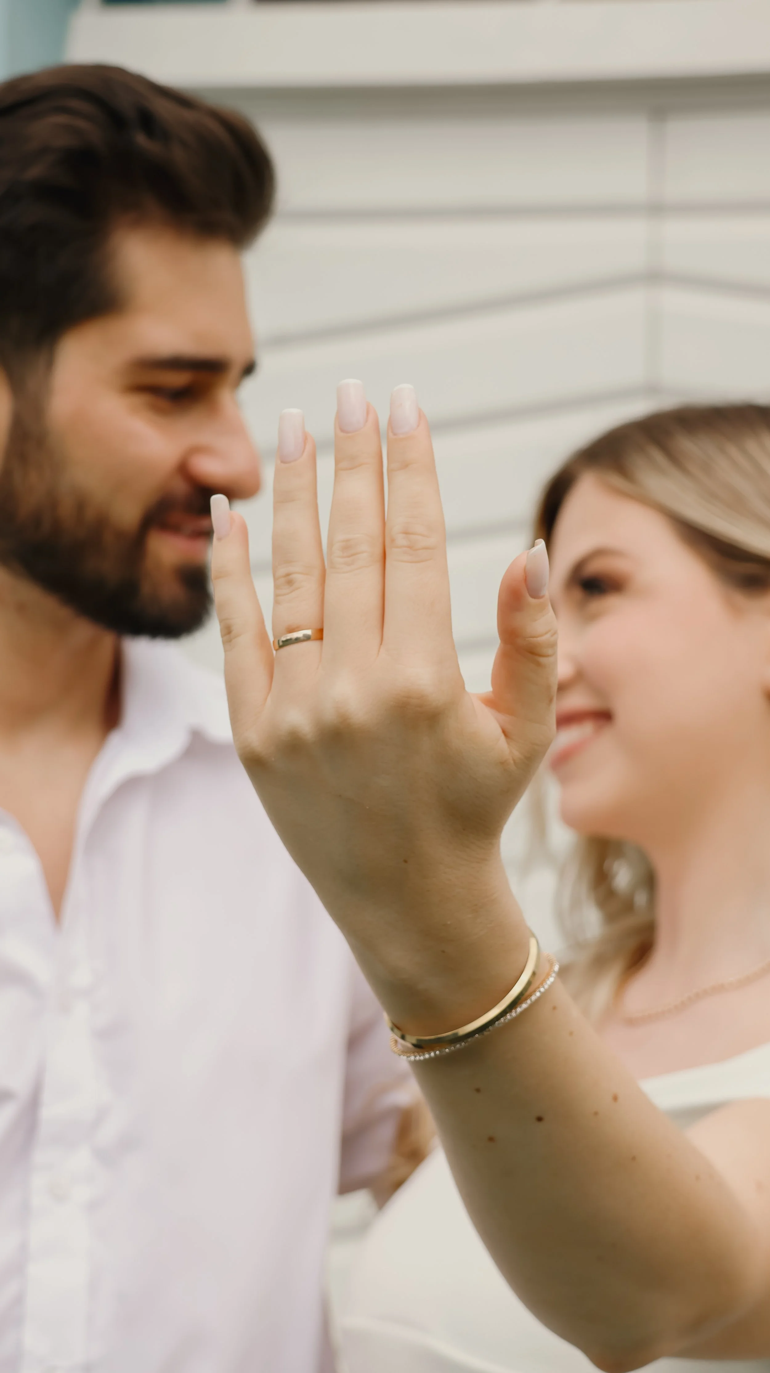 Um casal sorridente troca um beijo, com a mulher levantando a mão mostrando um anel de casamento, enquanto ela usa braceletes e tem unhas longas e bem cuidadas.