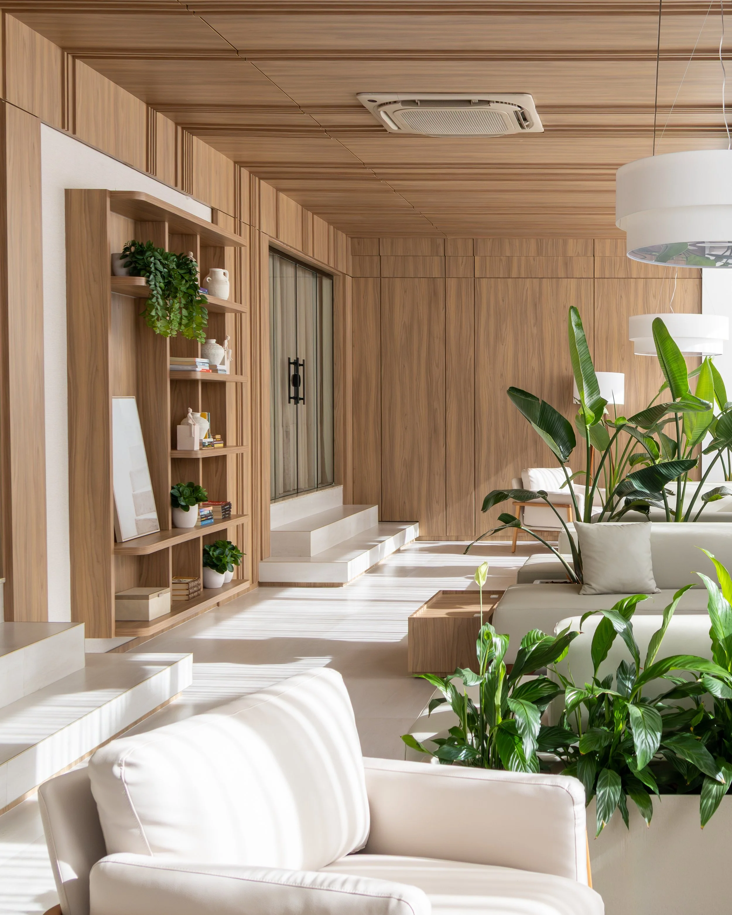 Sala de estar com móveis brancos, poltronas, plantas verdes, iluminação suave, paredes de madeira e prateleira de madeira decorada com livros e vasos.