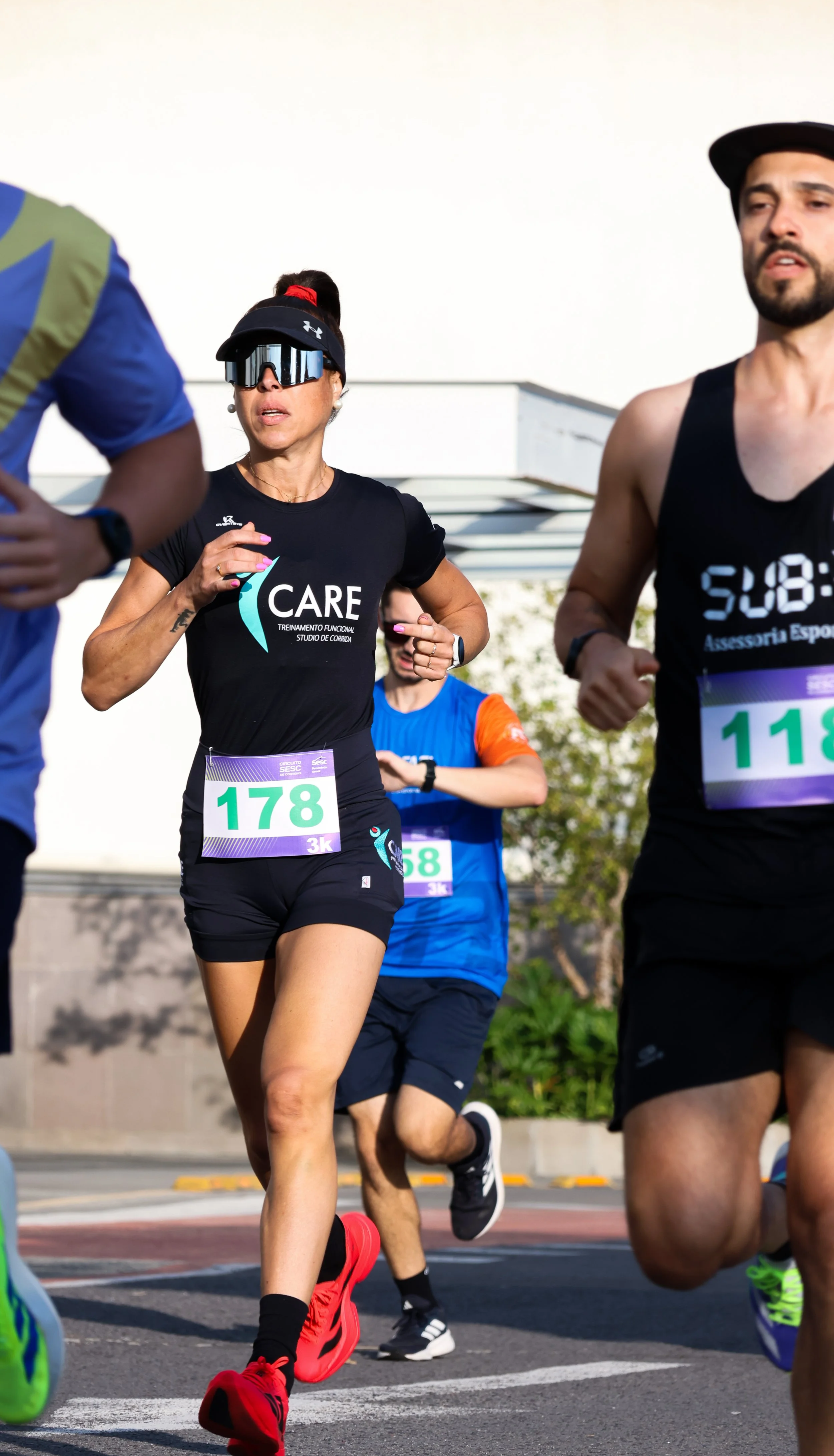 Corredores participando de uma corrida de rua, usando roupas esportivas e número de identificação. O foco é em uma mulher com óculos esportivos, camiseta preta e short preto, com o número 178.